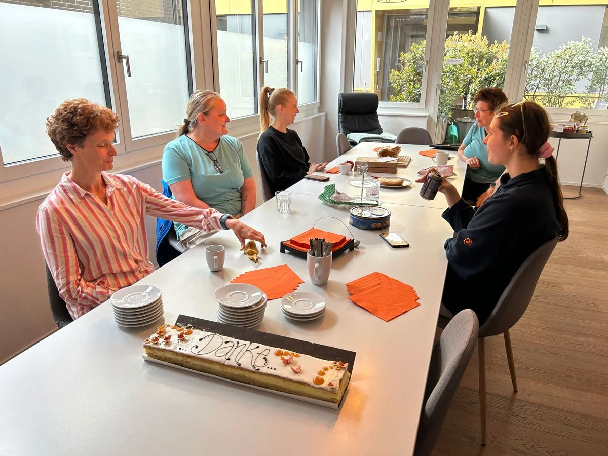 Fünf Frauen sitzen um einen weißen Tisch in einem Raum mit großen Fenstern. Auf dem Tisch liegen Teller, Tassen, Papierservietten, eine Geburtstagstorte mit "Danke" geschrieben, und andere Snacks.