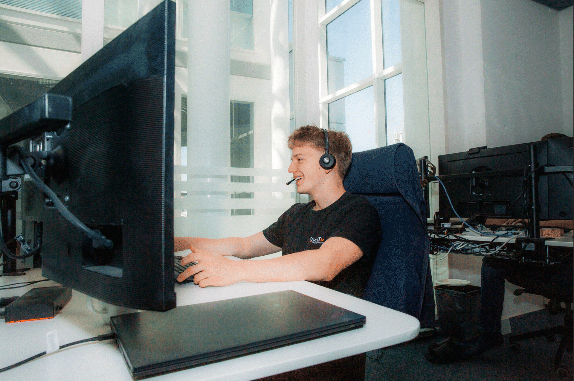 Junger Mann arbeitet an einem Computer mit mehreren Bildschirmen, trägt ein Headset und sitzt an einem Schreibtisch in einem modernen Büro.