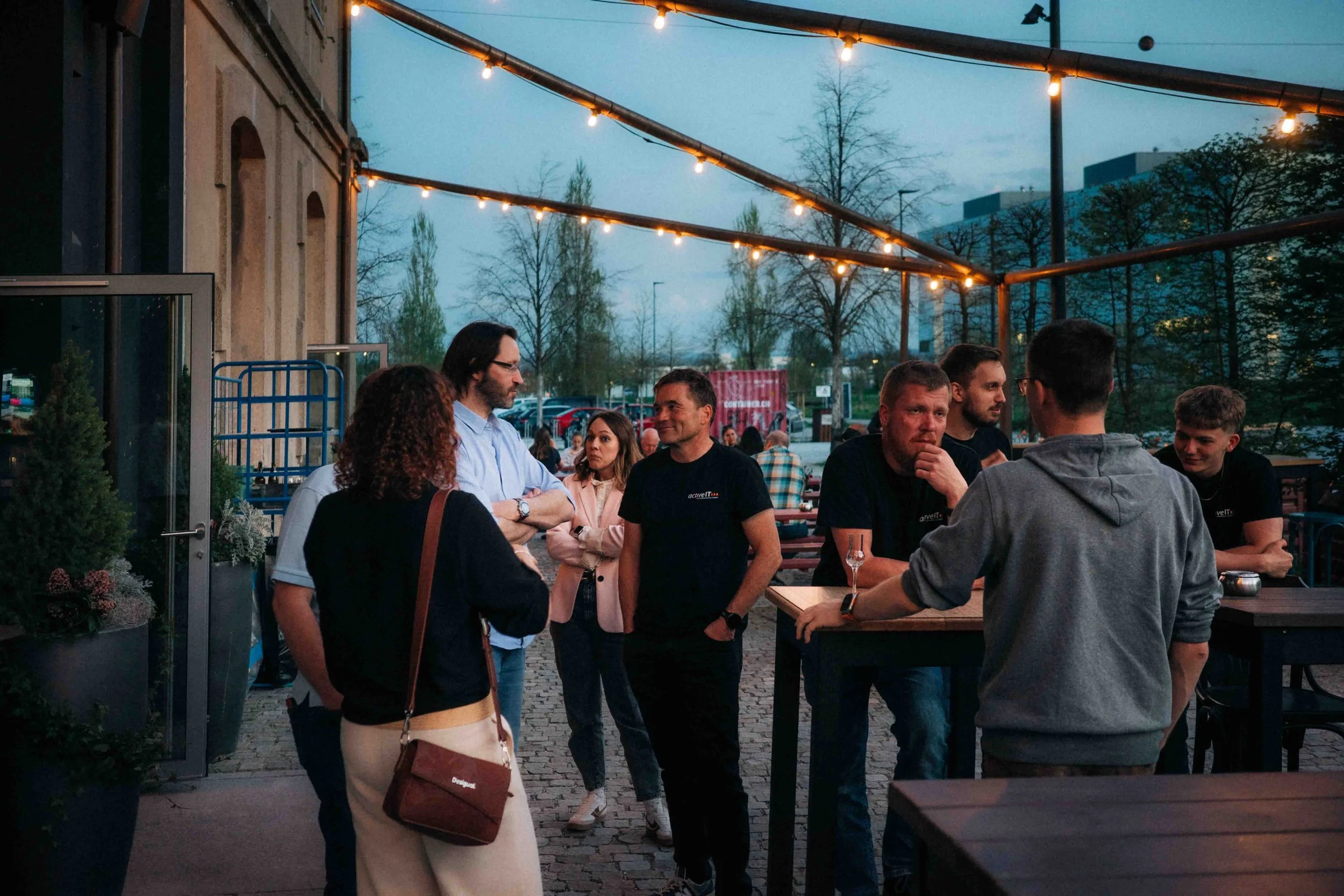 Menschen unter einer gemütlichen Outdoor-Lichterkette in einer Bar oder einem Restaurant im Freien bei Dämmerung oder Abend. Einige Menschen sprechen miteinander, einige trinken. Im Hintergrund sind Bäume und Gebäude sichtbar.