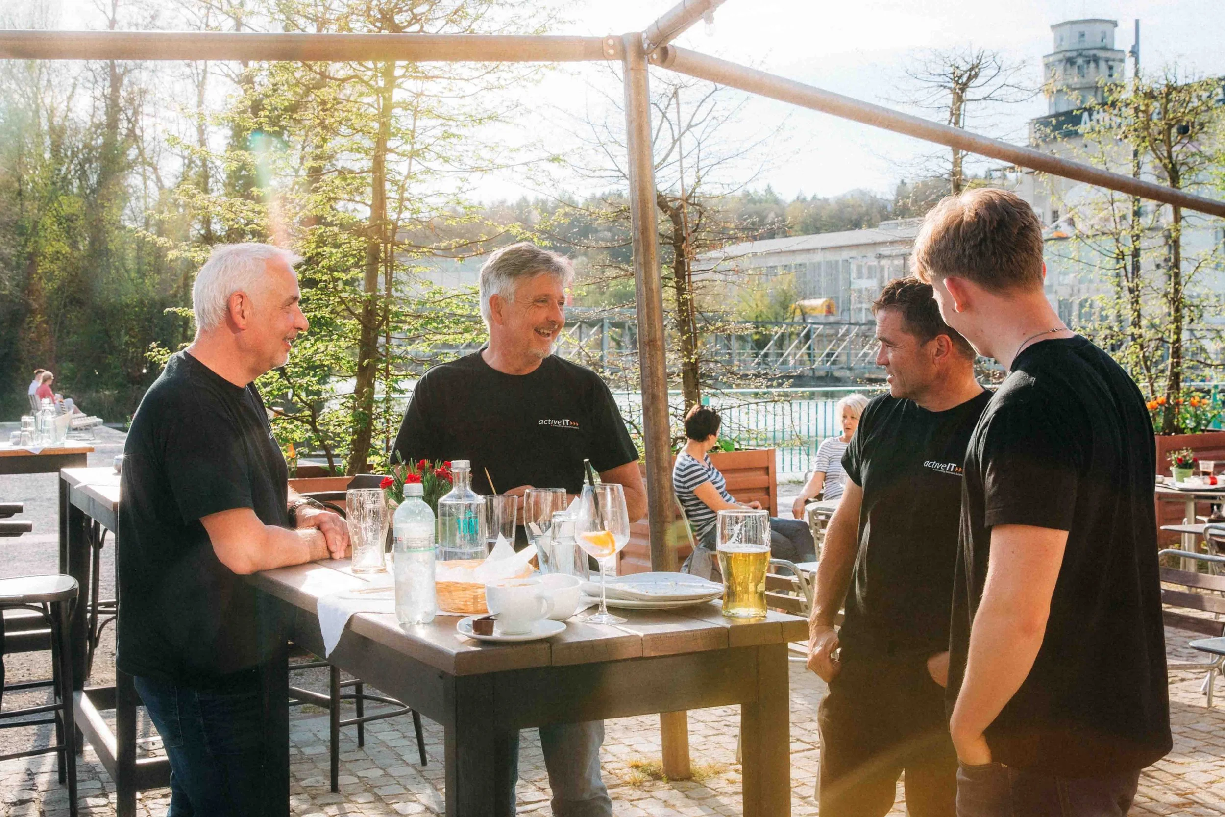 Vier Männer beim Gespräch an einem Tisch im Freien bei schönem Wetter, Getränke und Essen auf dem Tisch, im Hintergrund weitere Gäste und Bäume.
