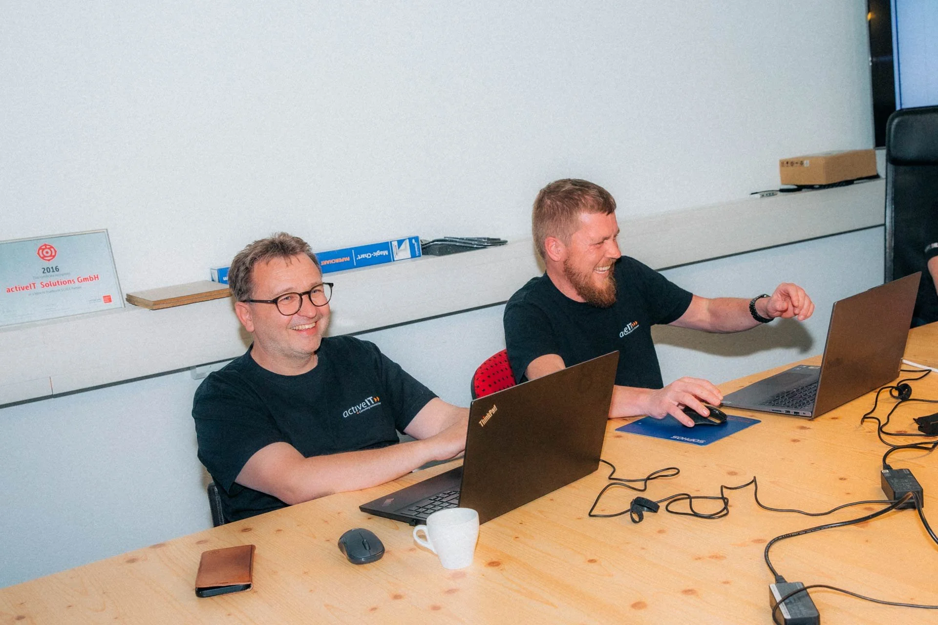 Zwei Männer lächeln bei einem Treffen in einem Büro. Sie sitzen an einem Holztisch mit Laptops. Einer trägt eine schwarze Brille, beide tragen schwarze T-Shirts mit einem Firmenlogo. Im Hintergrund befinden sich Bücher und eine weiße Tafel.