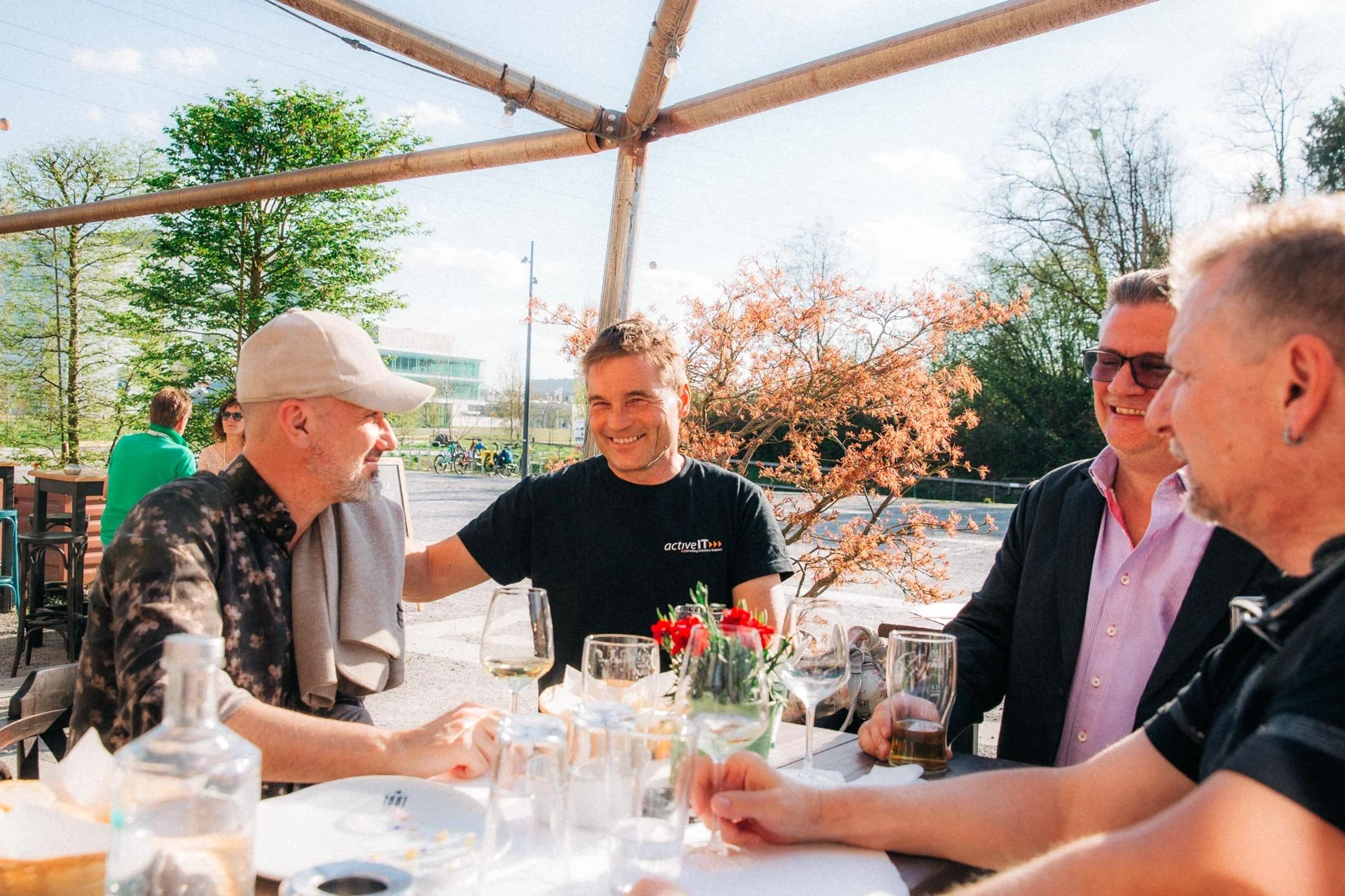 Eine Gruppe von Menschen sitzt bei einem Treffen im Freien an einem großen Tisch mit Getränken und Dekorationen bei schönem Wetter im Frühling oder Sommer.