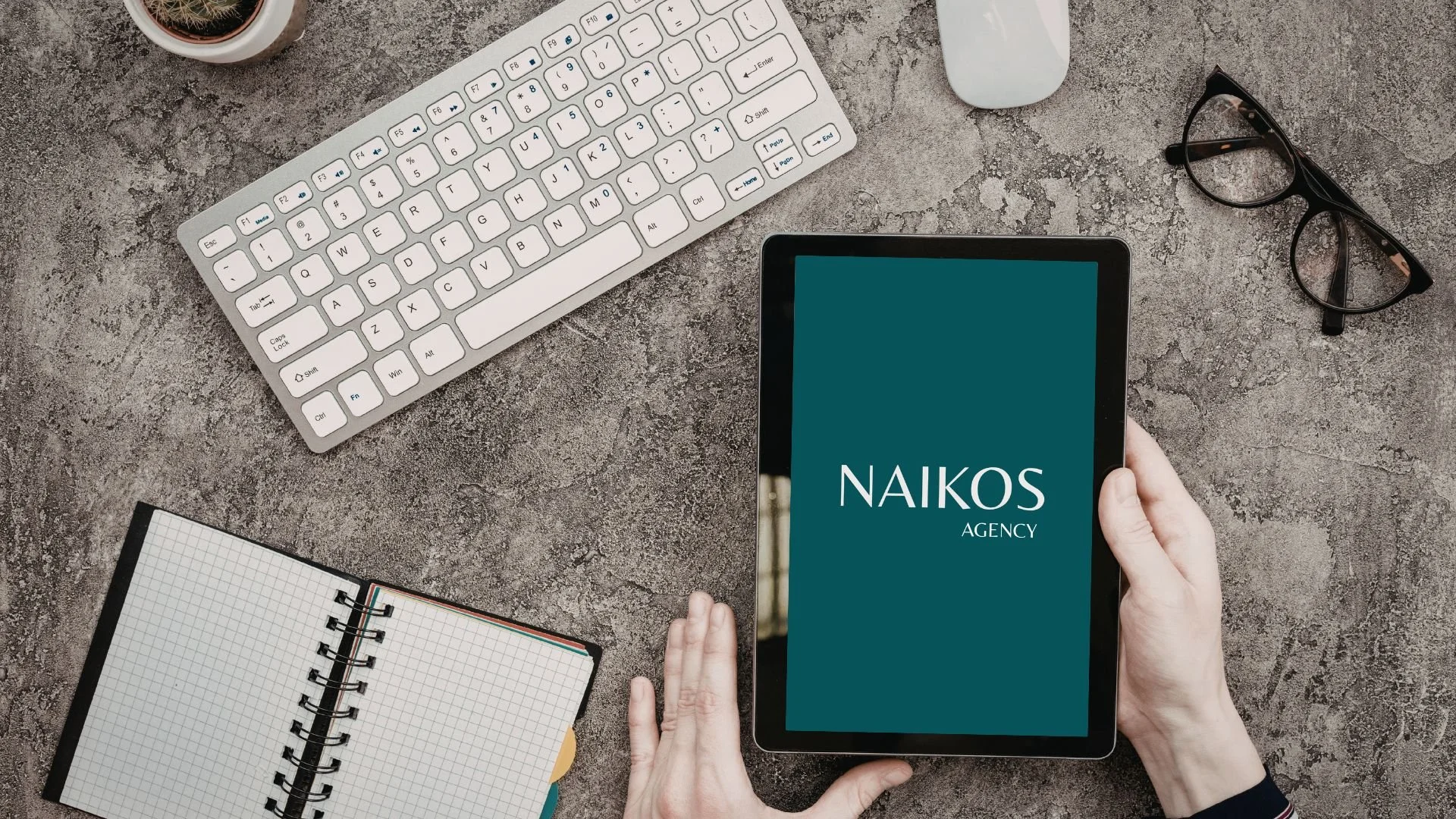 Vista de un escritorio con teclado blanco, gafas negras, una taza de café y una tablet que muestra el texto 'NAIKOS AGENCY'. Además, hay una libreta de dibujo con cuadrículas y un cuaderno con hojas en blanco.