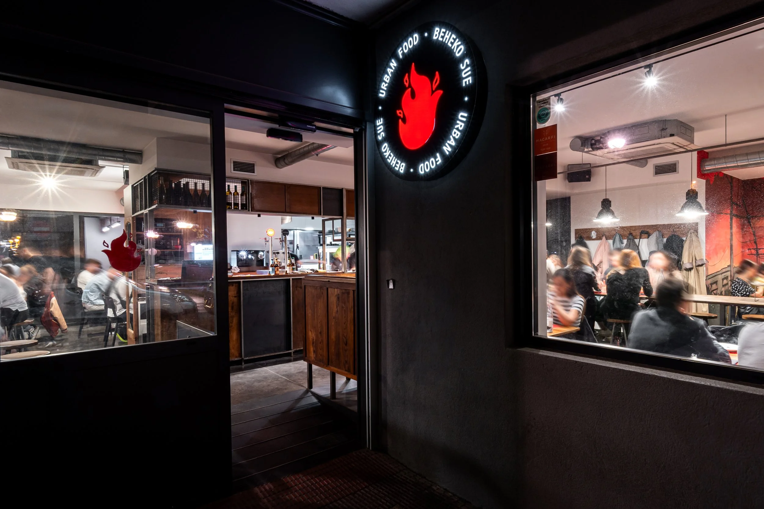 Vista de la fachada de un restaurante con ventanas transparentes, donde se observa el interior muy concurrido con muchas personas cenando. En la fachada hay un letrero circular con una llama en el centro y las palabras 'BEHEKO', 'URBAN FOOD', y similares.