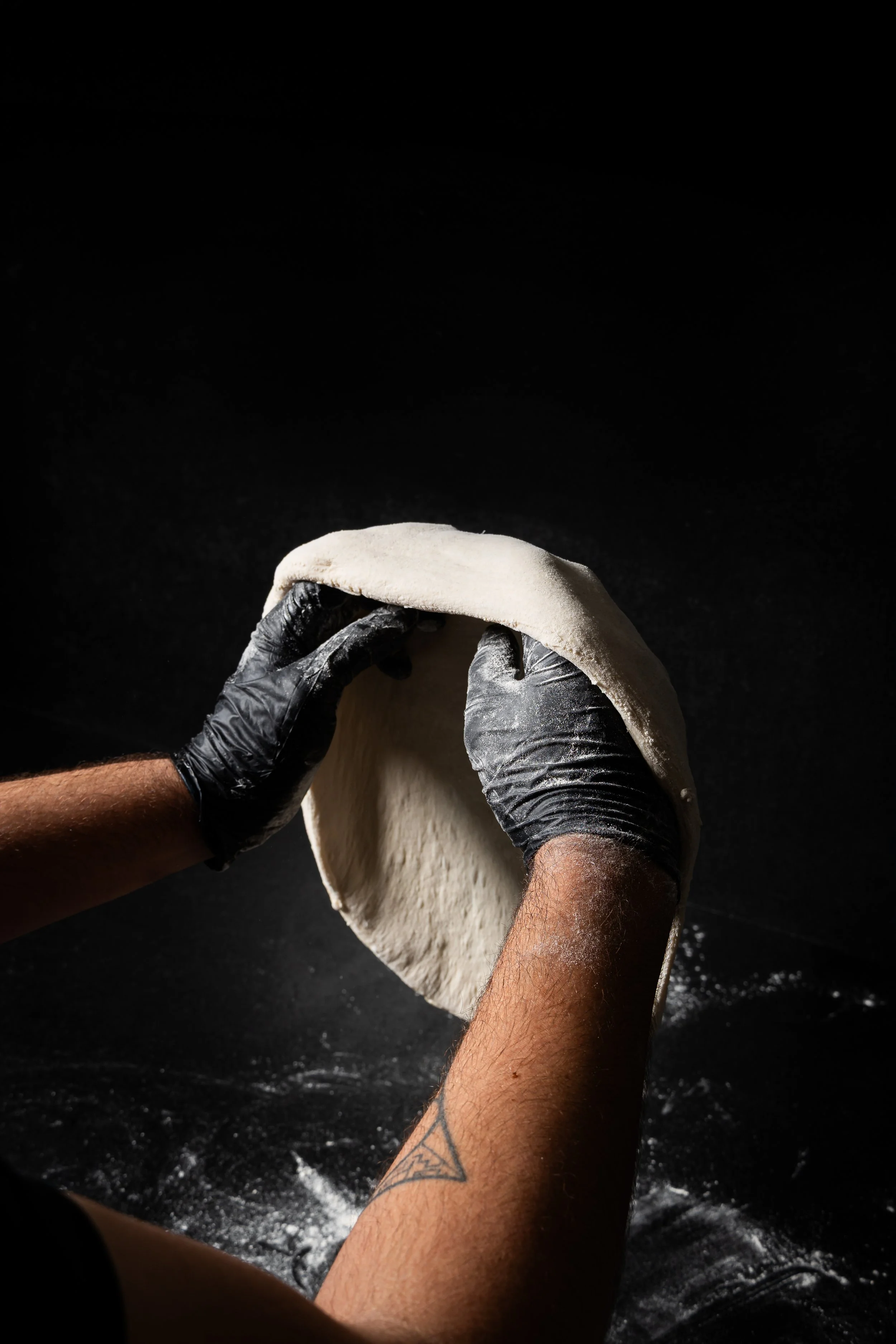 Personas haciendo pizza, usando guantes negros, con fondo negro y superficie de trabajo en la que se extiende la masa.