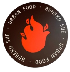Logotipo circular con una llama estilizada en color naranja en el centro y texto en ambos lados que dice 'BEHEKO SUE' y 'URBAN FOOD'.