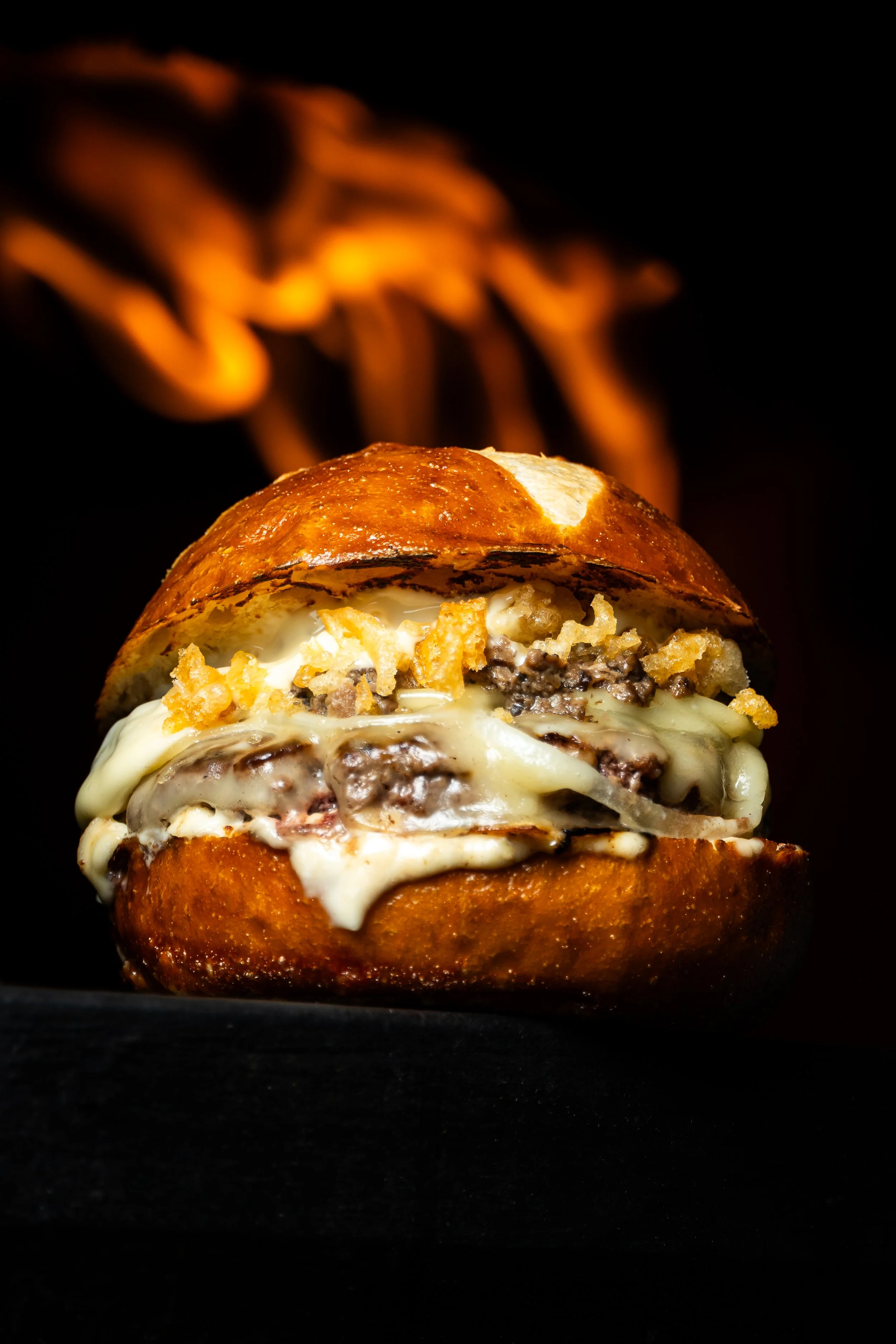 Hamburguesa con carne, queso, cebolla, y salsas, con fondo oscuro y fuego en el fondo.