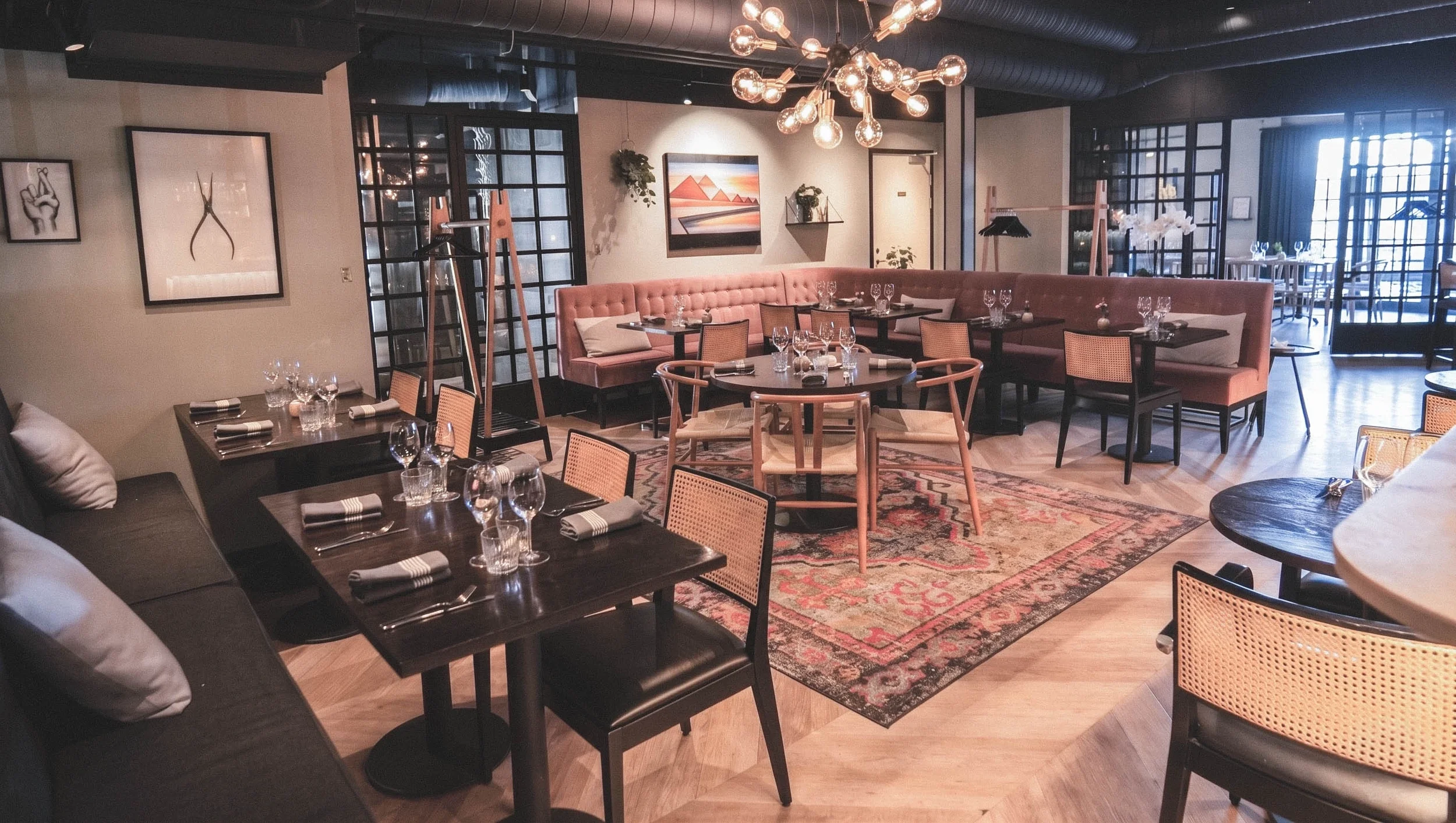 Ett modernt restauranginteriör med bord för middagsgäster, fåtöljer och stolar, väggdekorationer, ett stort ljuskrona i taket, och glasdörrar som leder till en utomhusveranda.