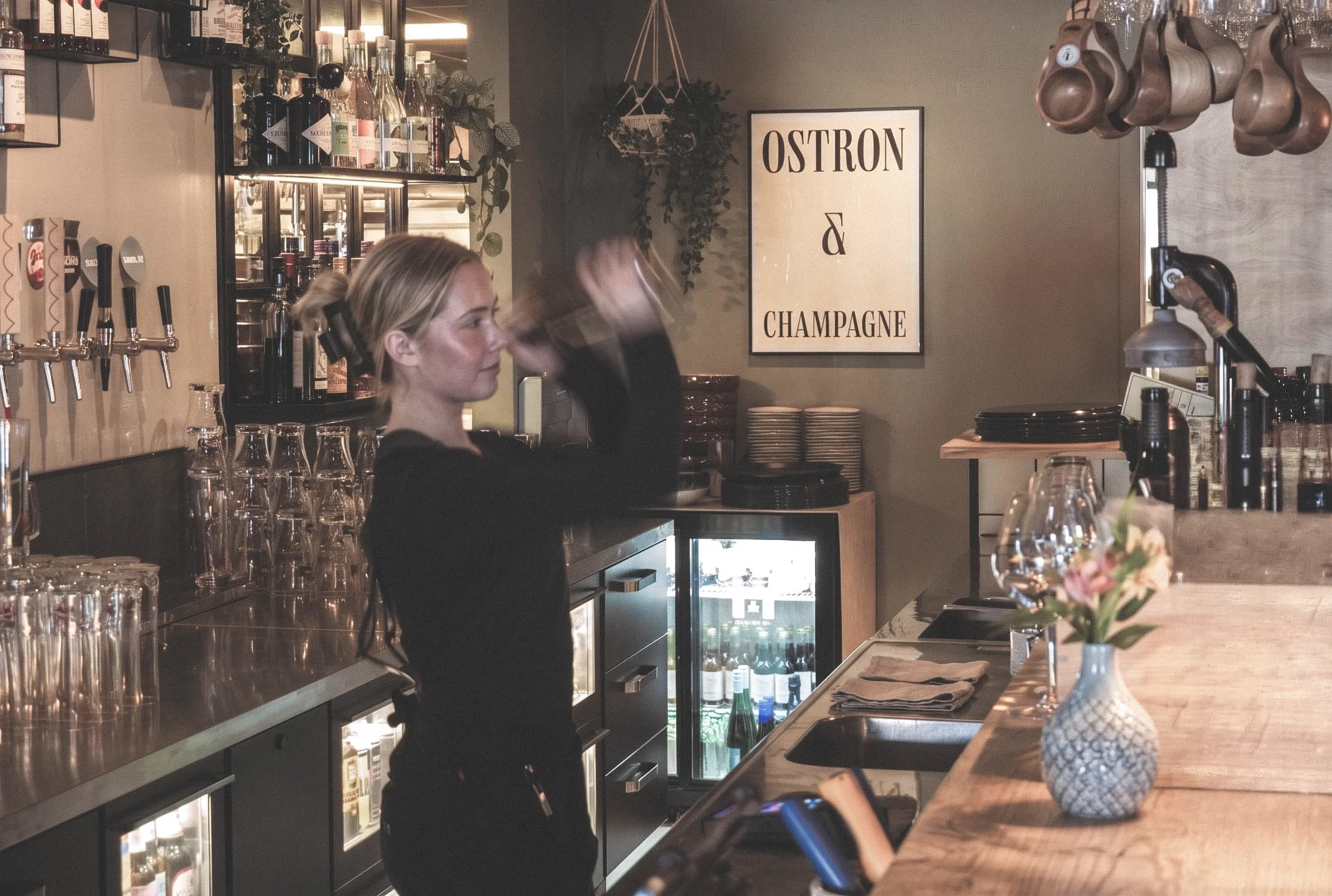 En kvinna som står bakom baren i en bar eller restaurang, fyller ett glas med dryck. Bakom henne finns hyllor med flaskor och glas.