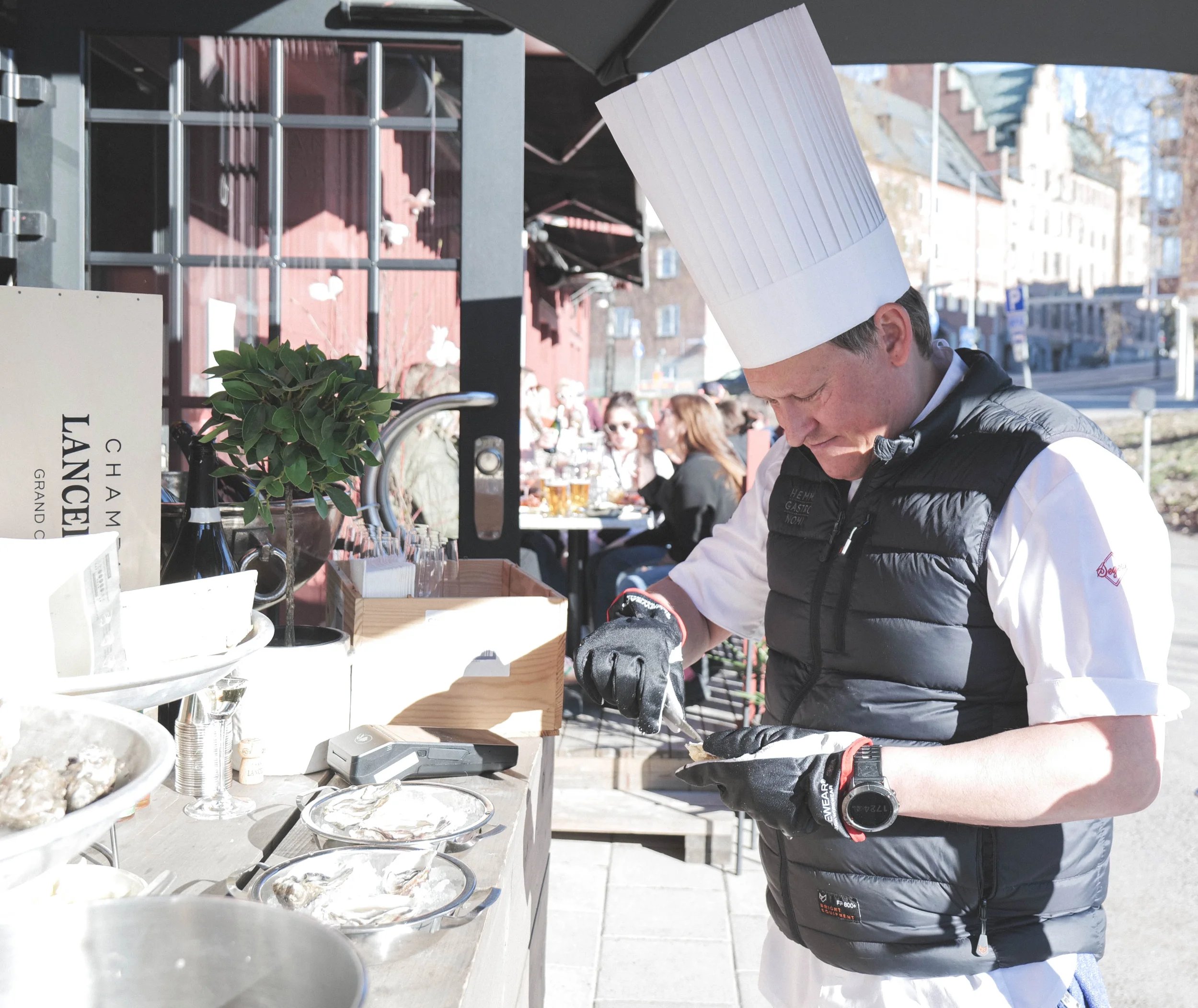 Sinom Laiti  i svart förkläde och vit kockhatt arbetar vid en matstång utomhus, med flera serveringsfat med mat och drycker framför sig. Det finns en växt och en skylt med texten 'Champagne' i förgrunden.