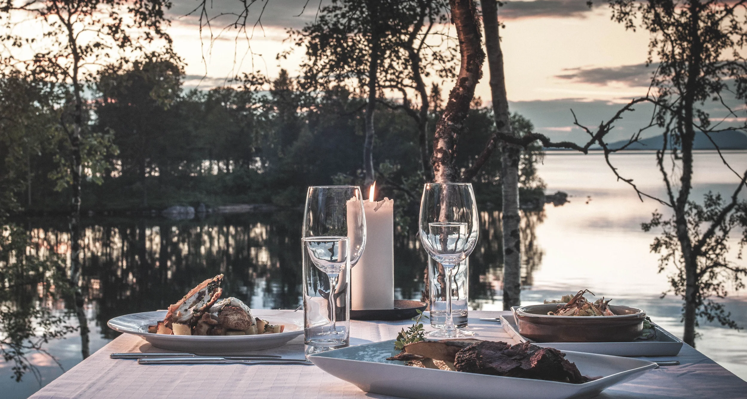 En middagsscen med mat, två tomma vinkoppar och ett ljus framför en sjö vid solnedgången. Trestammar och lövträd syns i bakgrunden.