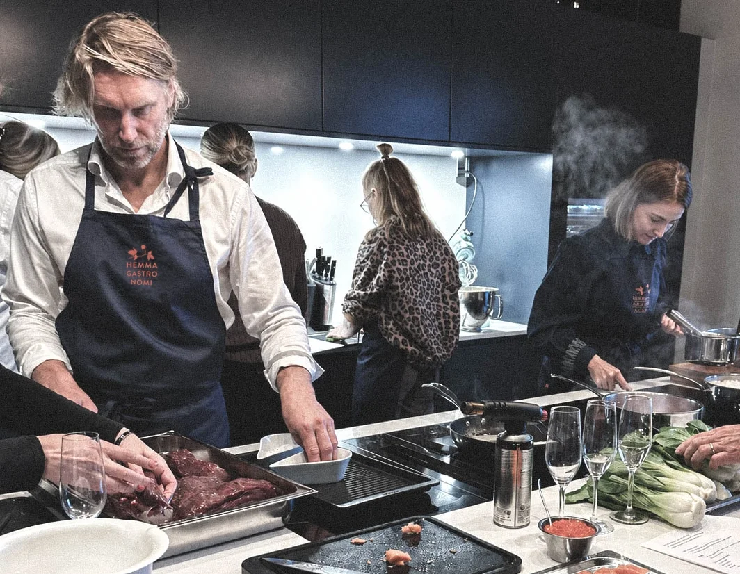 Människor som lagar mat i ett kök, inklusive en man som hanterar kött och en kvinna som steker något på spisen.