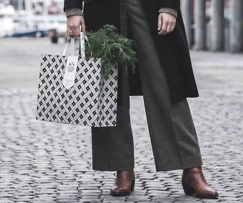En person som bär svarta byxor och bruna stövlar, håller i en Hemmagastronipåse med grön dill i, står på en kullerstensgata i stadsmiljö.