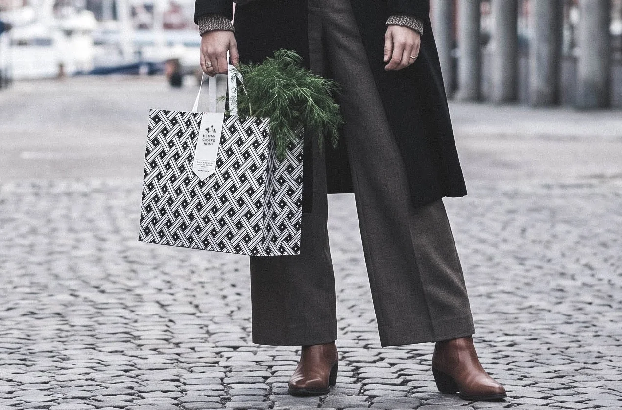 En person som bär svarta byxor och bruna stövlar, håller i en Hemmagastronipåse med grön dill i, står på en kullerstensgata i stadsmiljö.