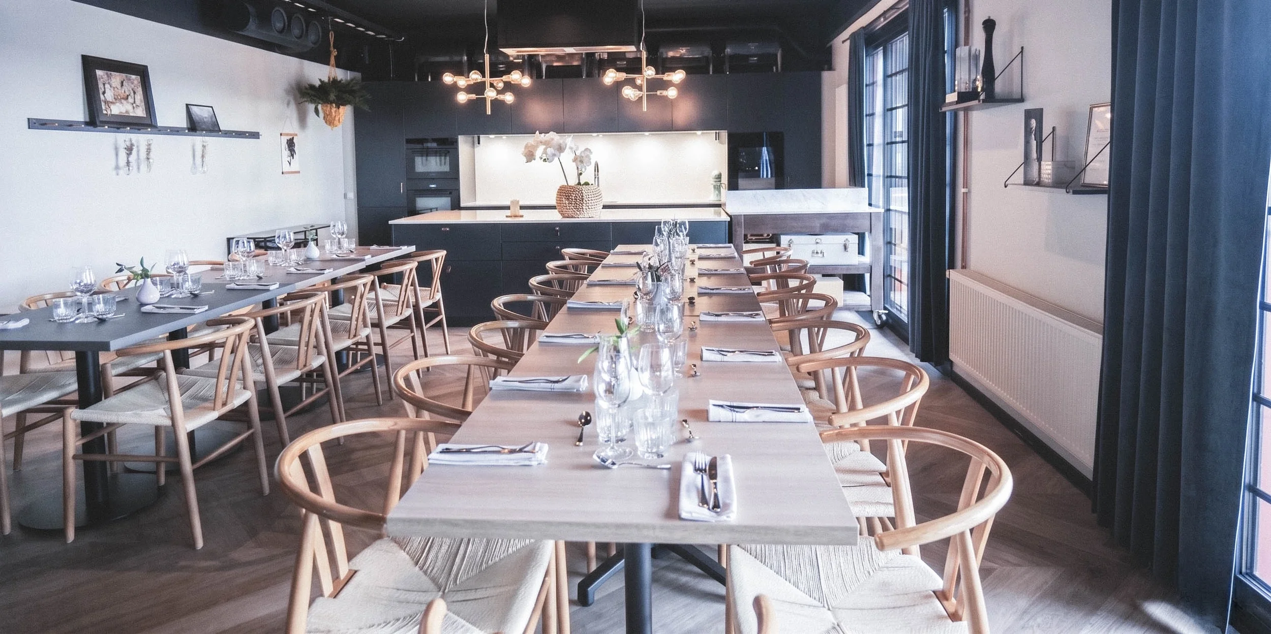 Ett tomt matsalsrum med rad av dukade bord, glas, bestick och servetter. Moderna möbler och väggdekorationer syns, samt fönster med mörka gardiner.