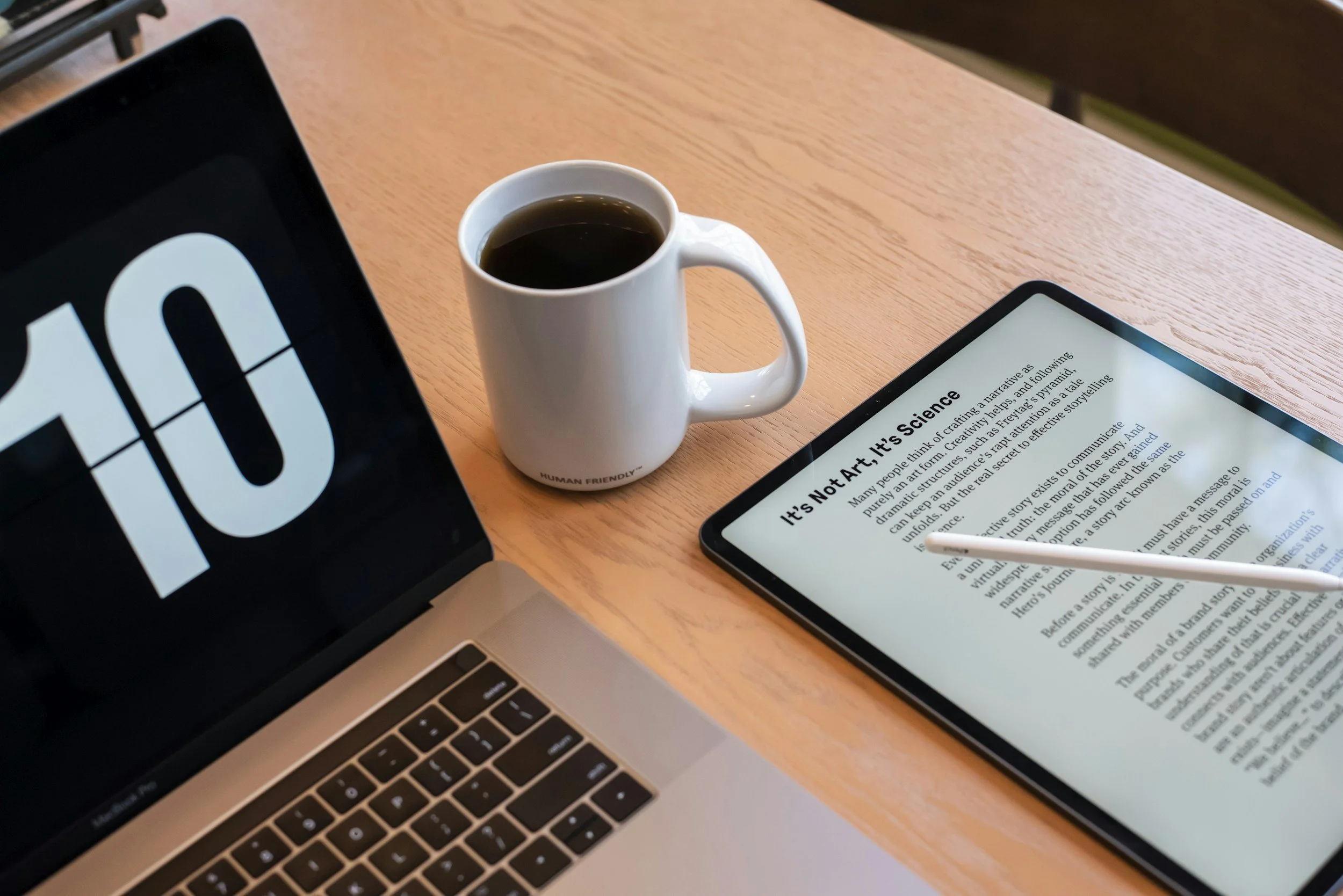 Un ordinateur portable, une tablette avec un stylet, une tasse de café noir, posé sur une table en bois clair.