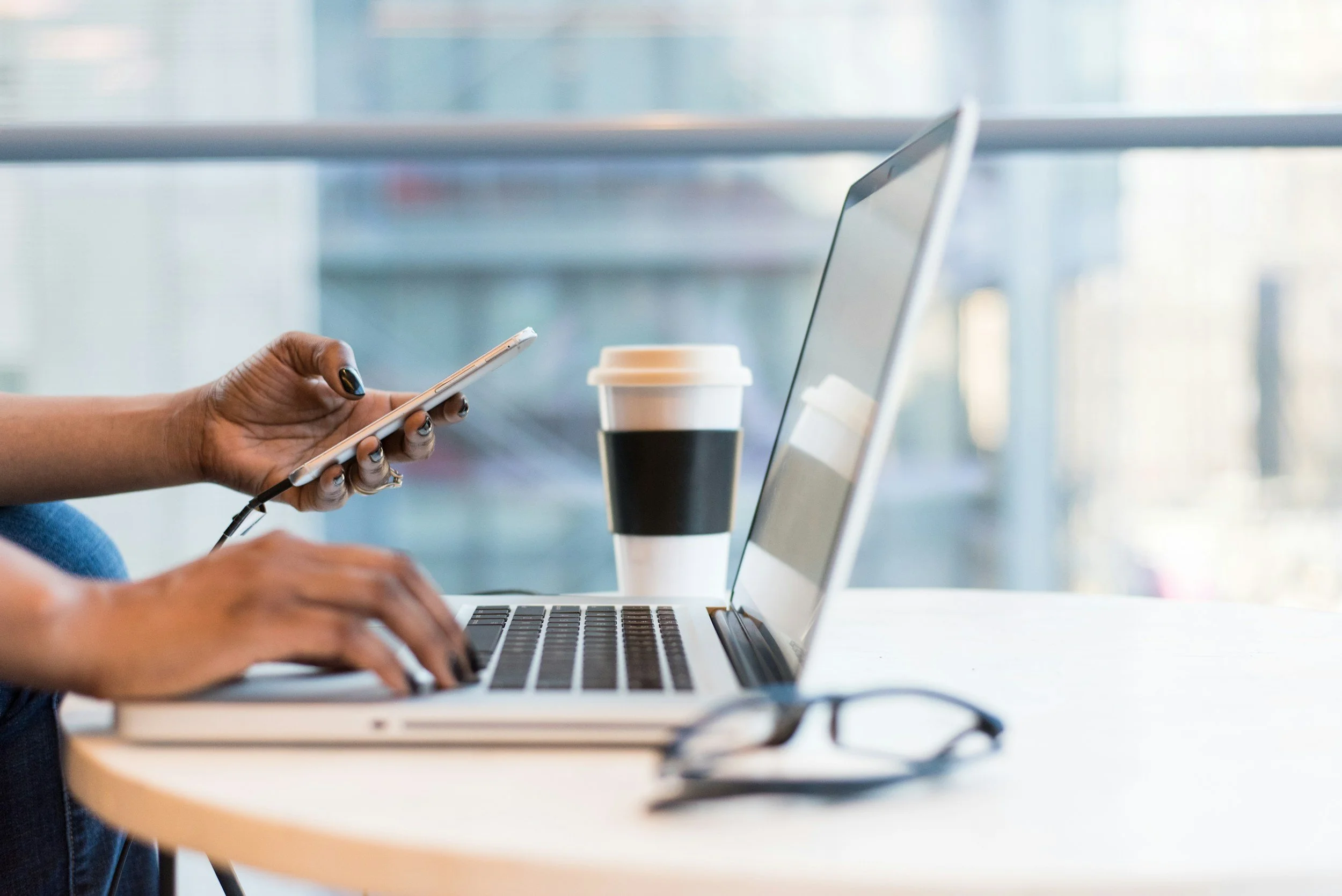 Une personne utilisant un smartphone avec un ordinateur portable et un café dans un environnement intérieur lumineux.