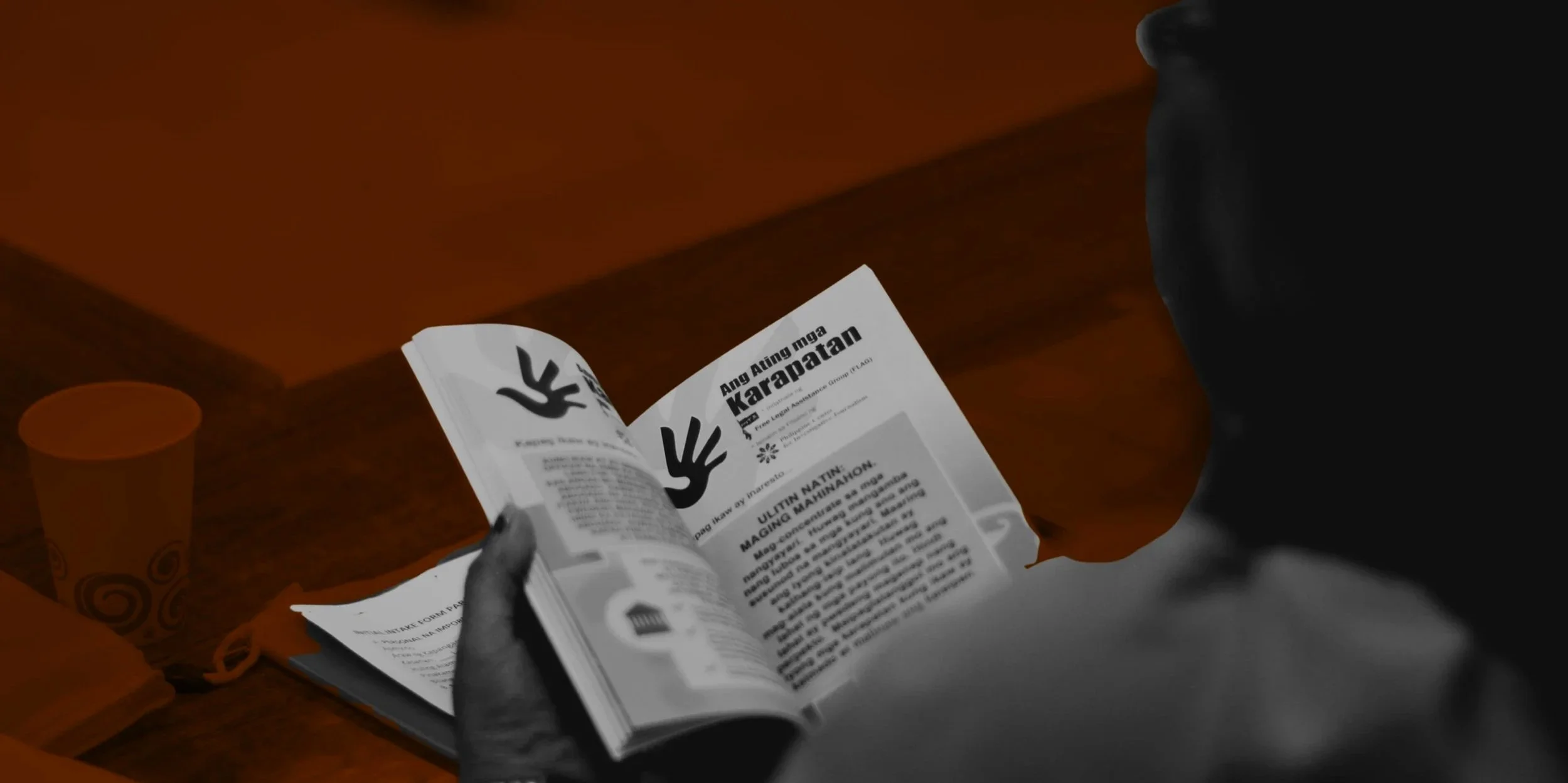 Person reading a magazine or brochure on a wooden table, with a cup nearby.