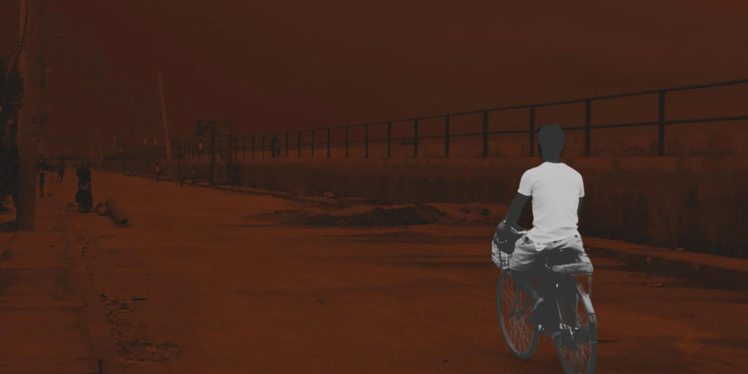A person riding a bicycle along a street during sunset or twilight, with a metal fence and a few distant figures in the background.