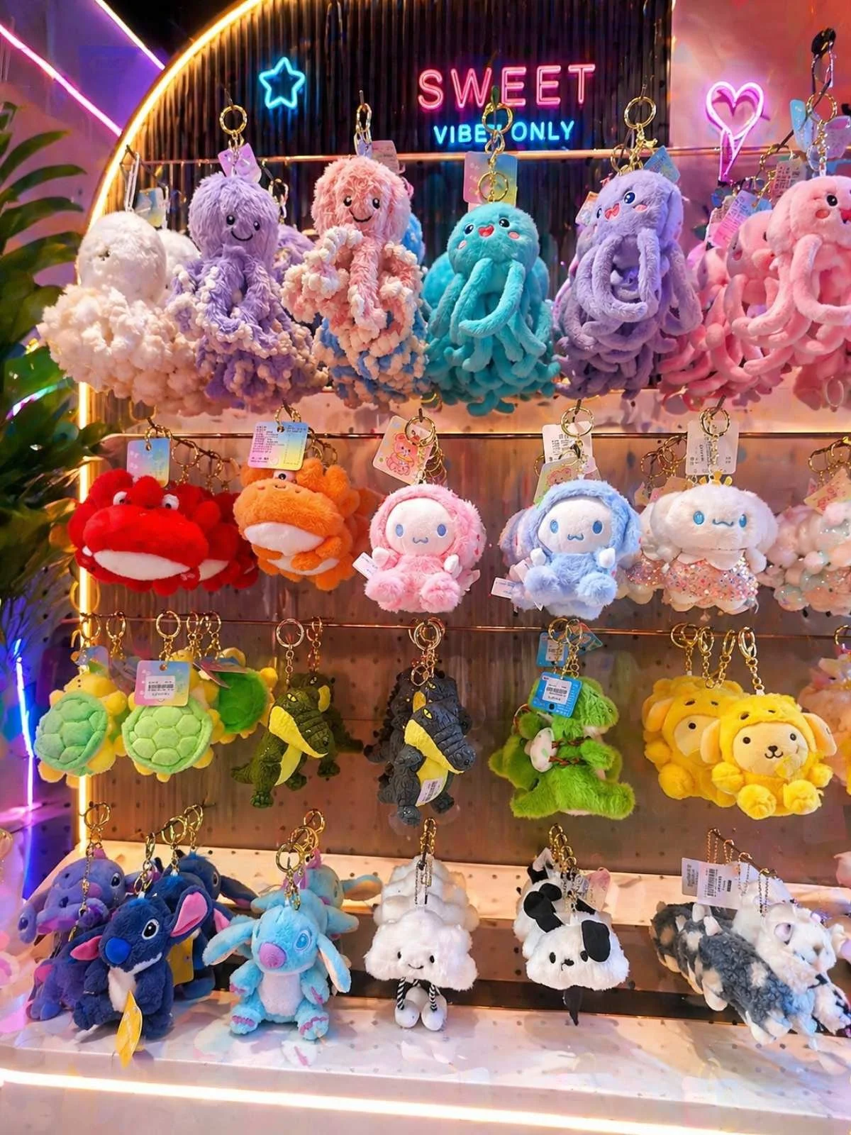 Colorful assortment of plush keychains shaped like animals and characters on display at a store. The background features neon signs reading "SWEET VIBE ONLY" and decorative neon shapes, including hearts and stars.