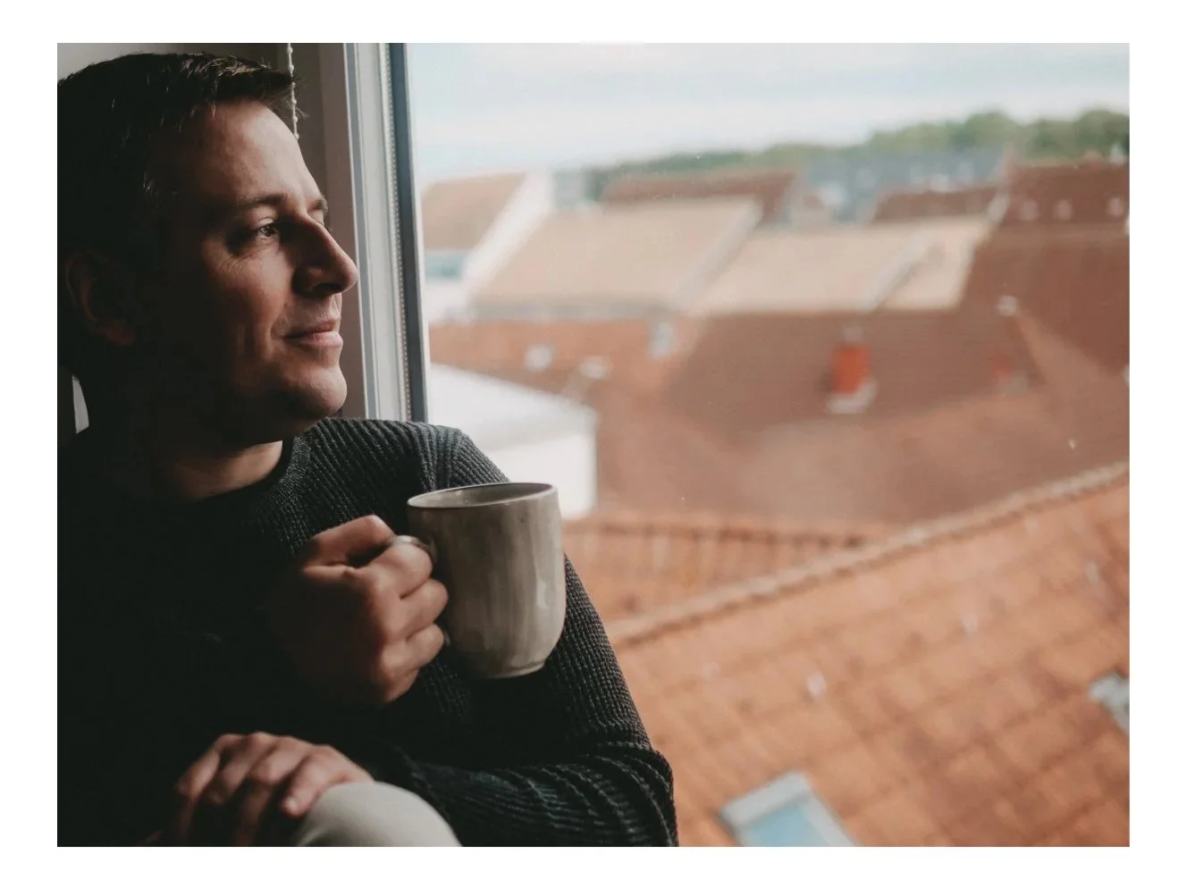 Mann sitzt am Fenster mit einer Tasse Kaffee und blickt nach außen, im Hintergrund rot-blaue Dachflächen.