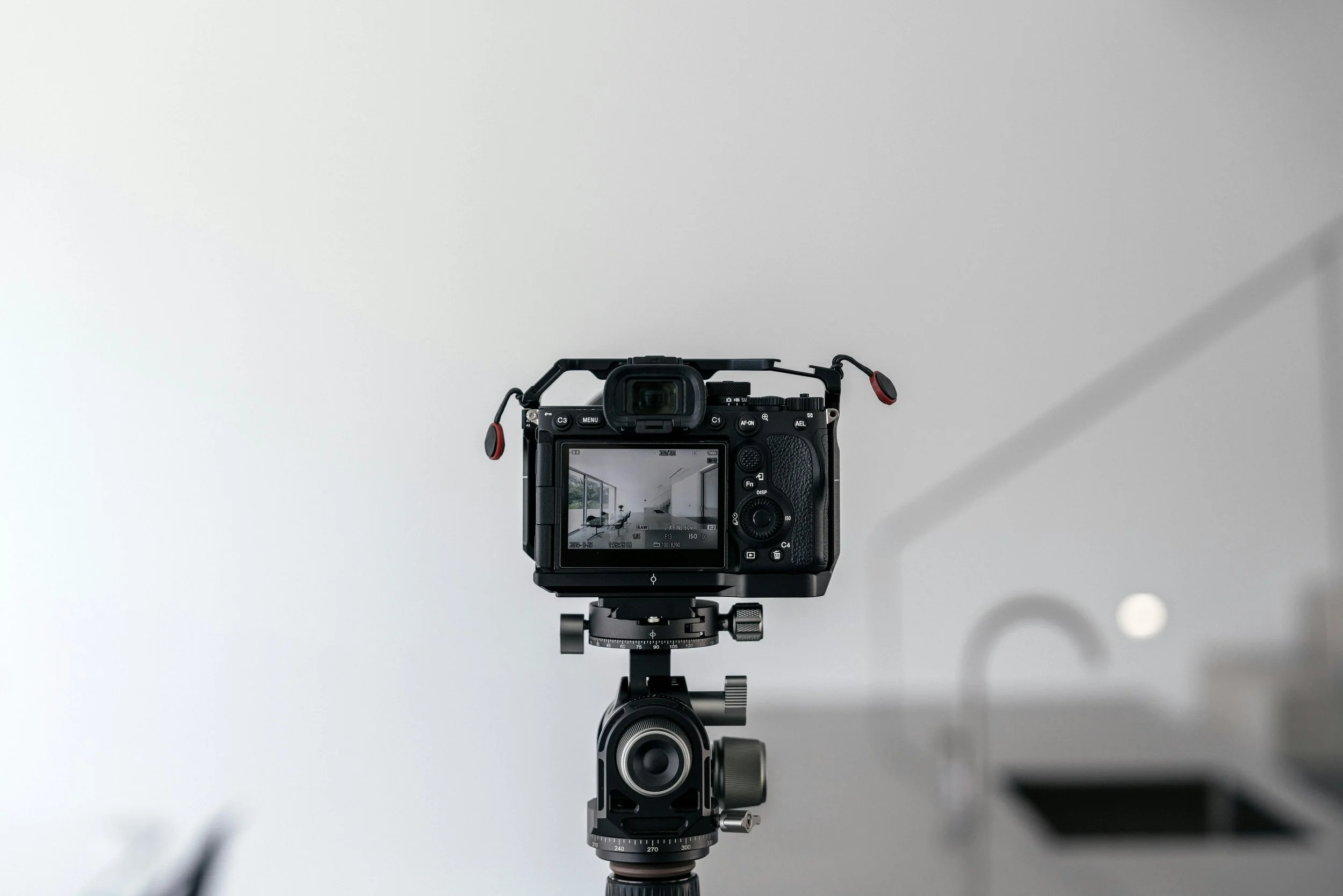 Camera mounted on a tripod positioned against a plain white wall, with a view of the camera's LCD screen showing a room with large windows and a sofa.