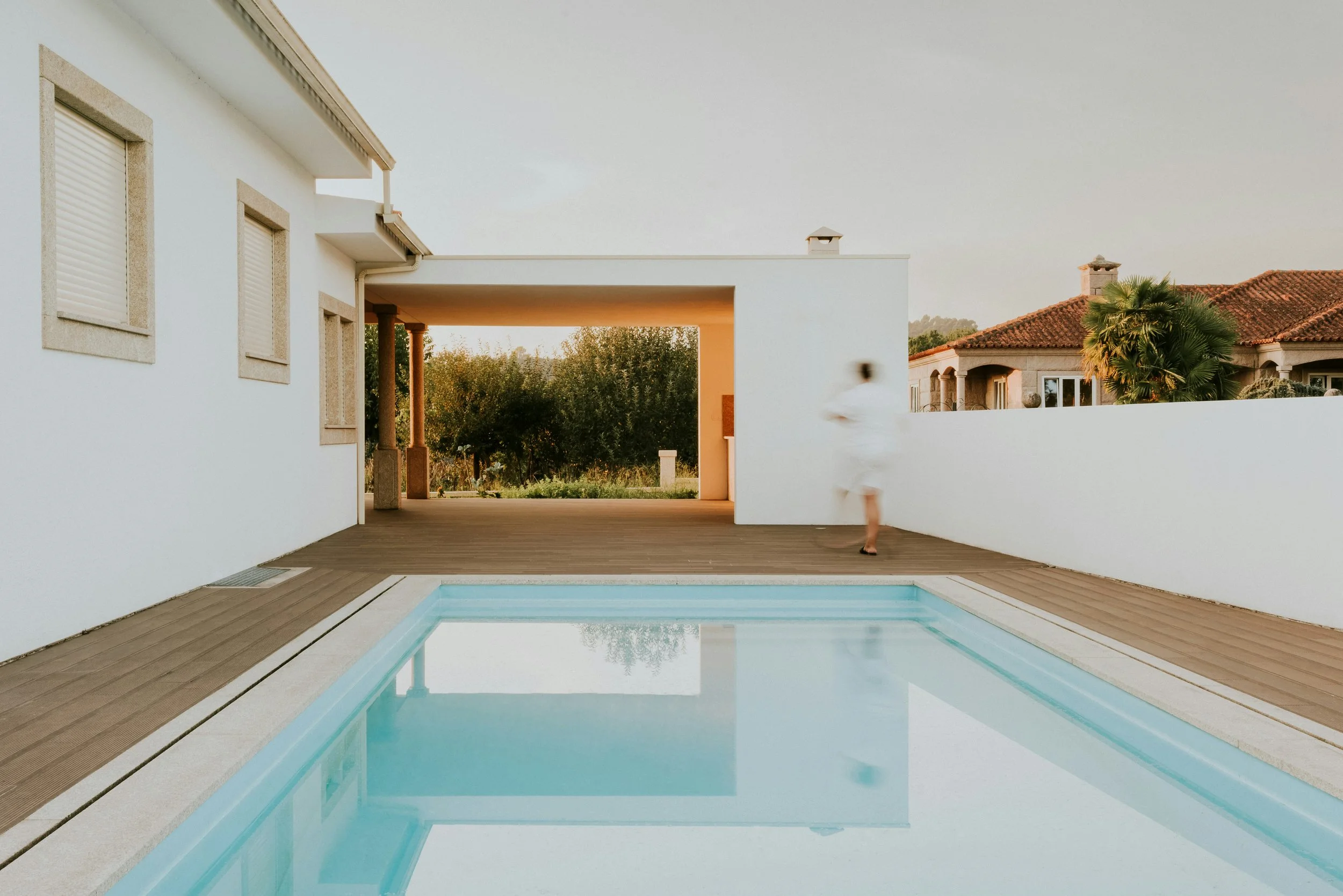 Modern house with a swimming pool in the backyard, surrounded by palm trees and a wooden deck, during sunset.