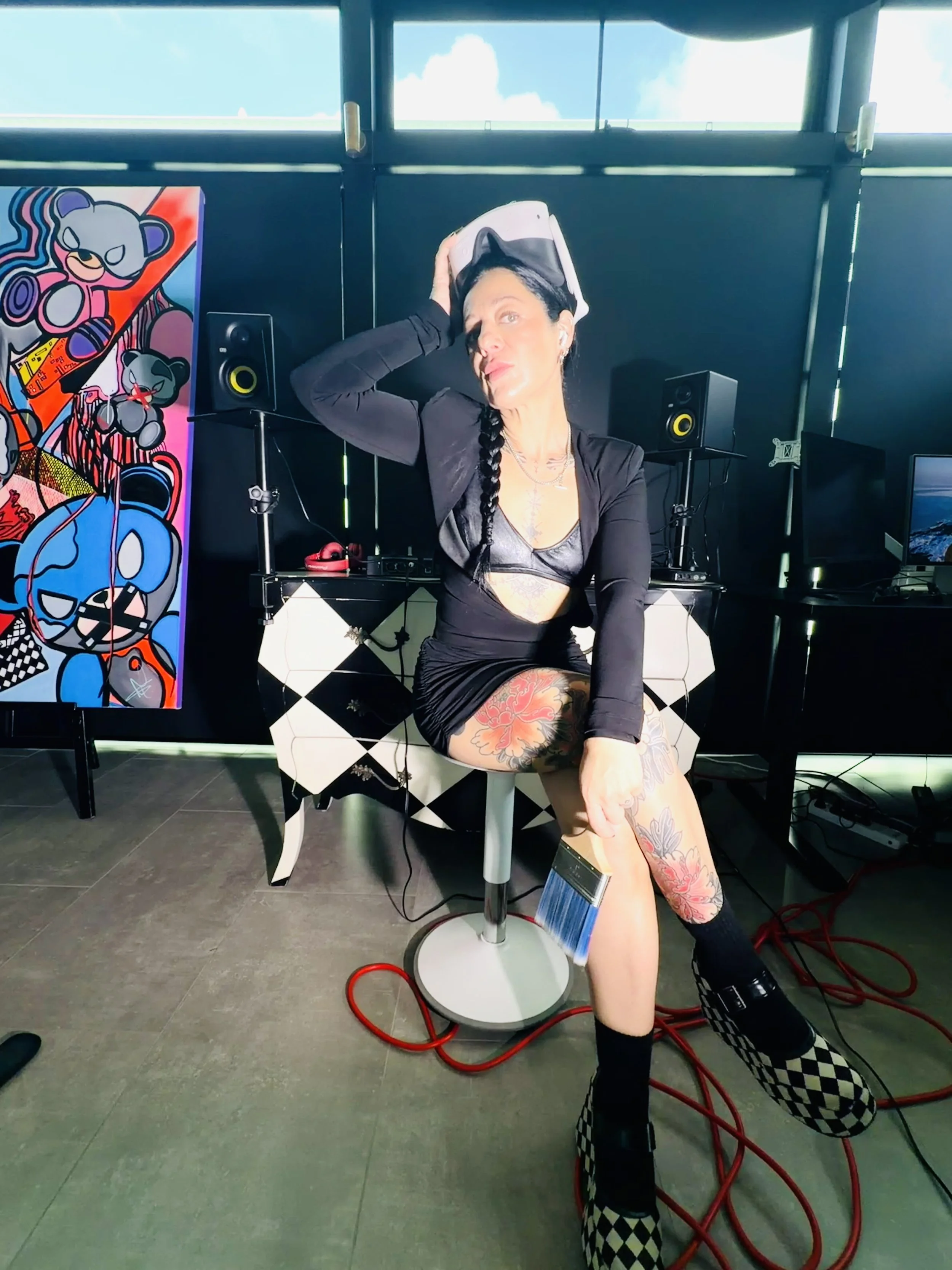 A woman with tattoos on her legs wearing a black crop top, a black jacket, and checkered platform boots, sitting on a chair, holding a virtual reality headset on her head in a room with DJ equipment, speakers, colorful artwork of cartoon bears, and a computer, with cloudy sky visible through windows.
