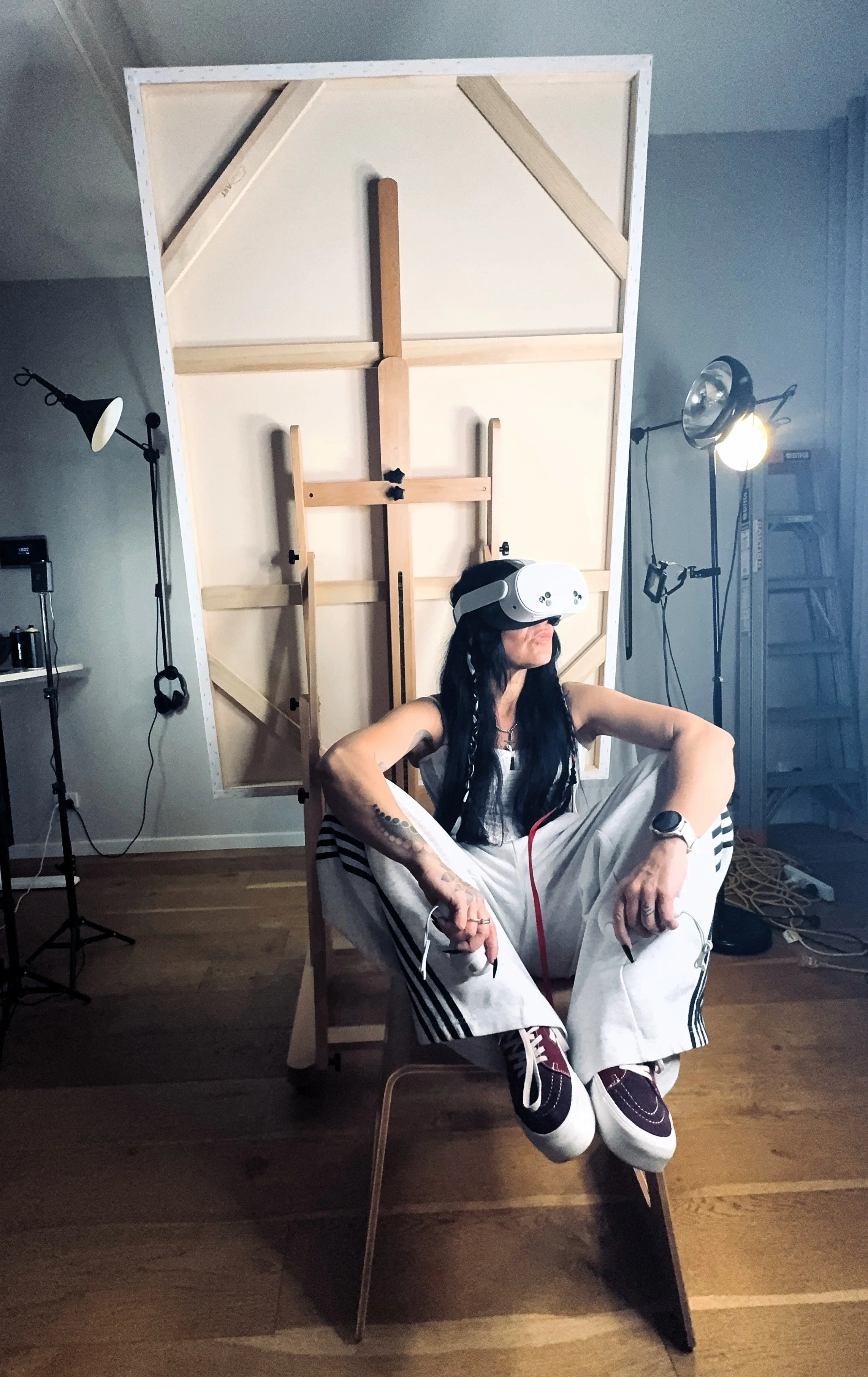 Person sitting on a chair wearing a virtual reality headset, surrounded by studio lights and equipment, with a large wooden art frame or installation behind her.