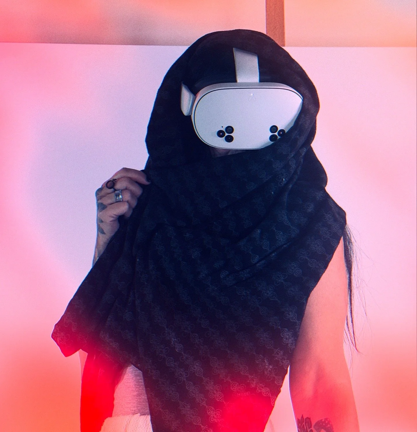 Person holding a scarf over the lower face, wearing a virtual reality headset, black scarf, dark clothing, and tattoos on the arm, with pink and red gradient background.