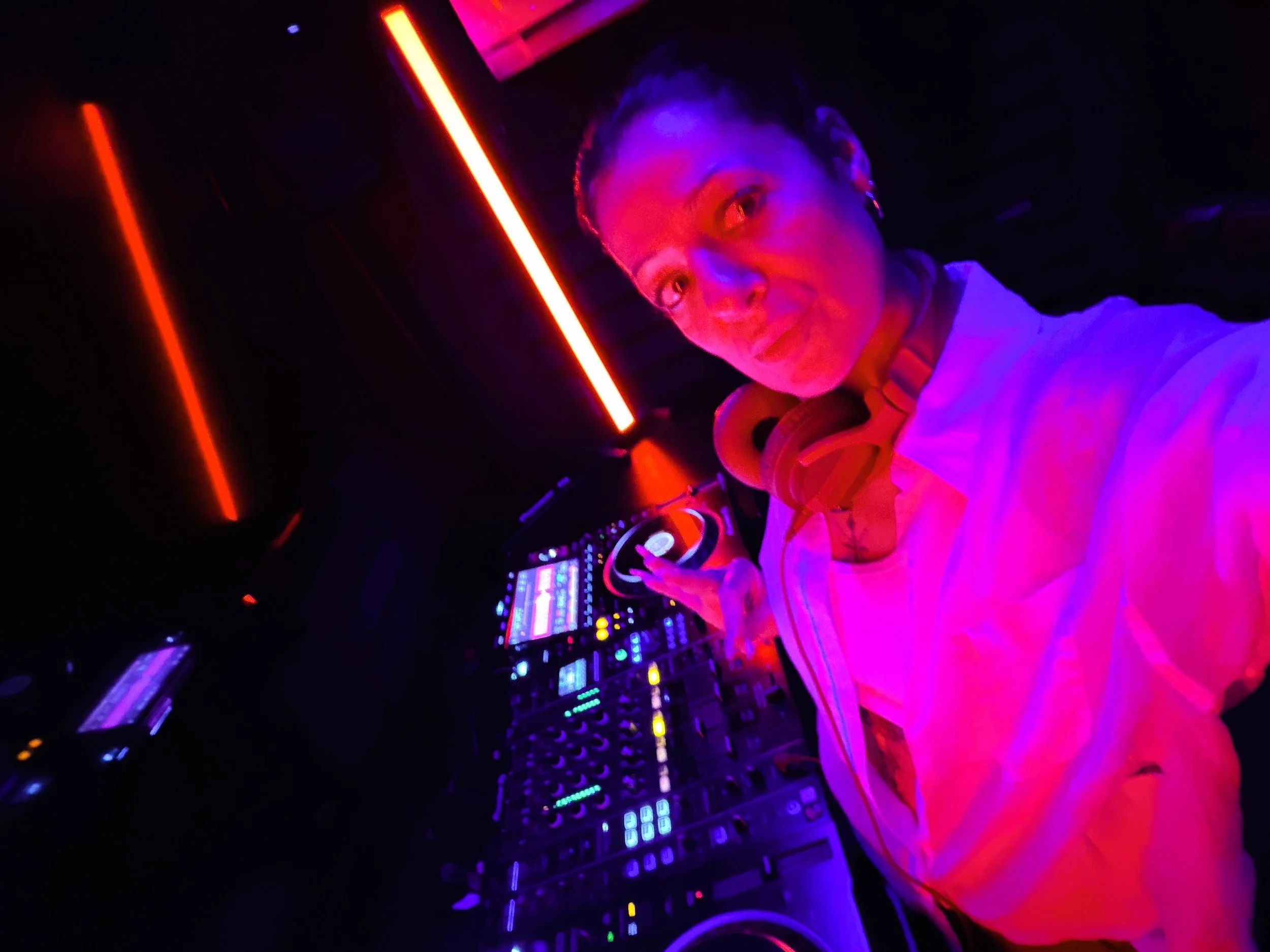 A person with headphones in a dark room with neon purple and orange lighting, standing in front of a DJ controller or mixing console with various illuminated controls and screens.