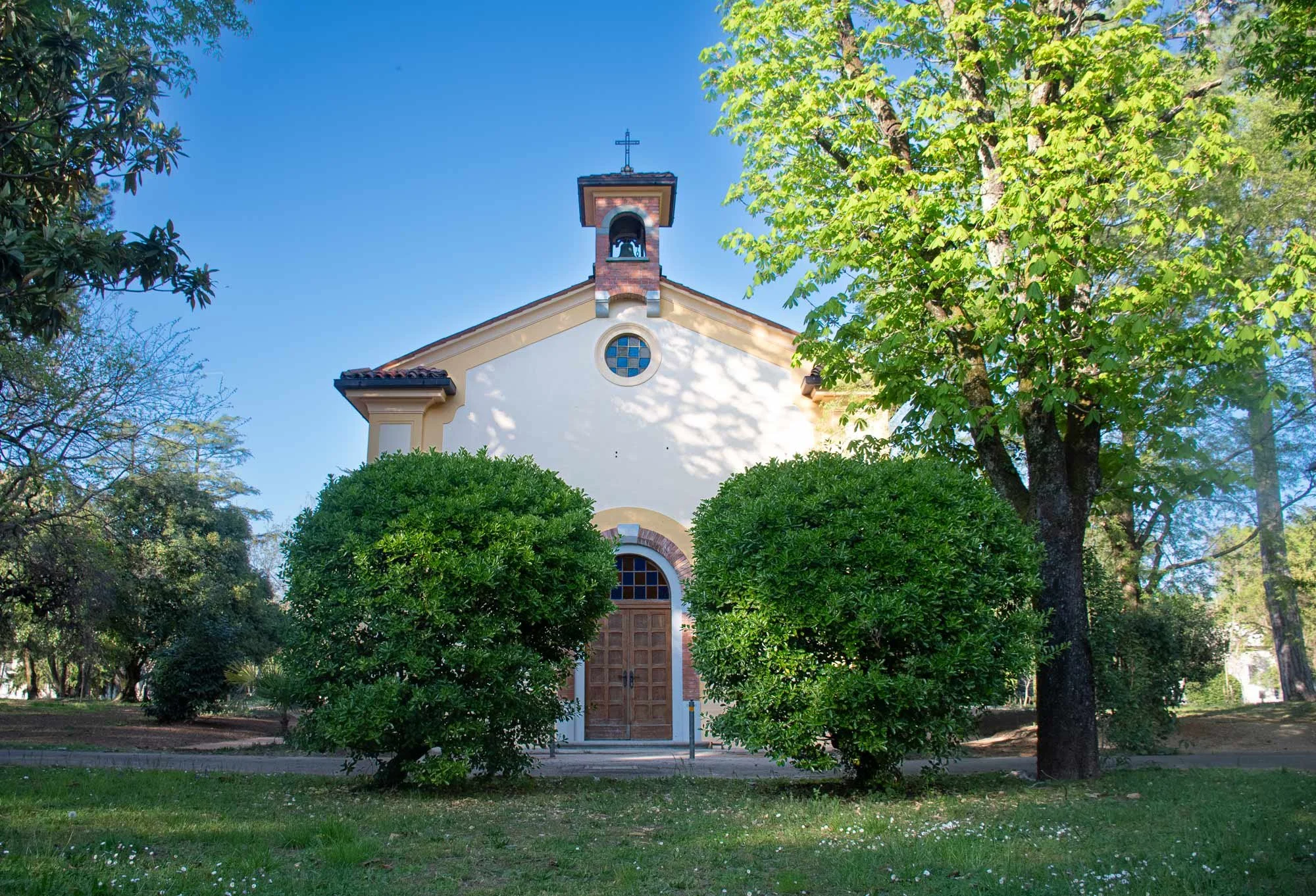 Chiesa bianca con portale di legno, circondata da alberi verdi e un prato, sotto un cielo azzurro.