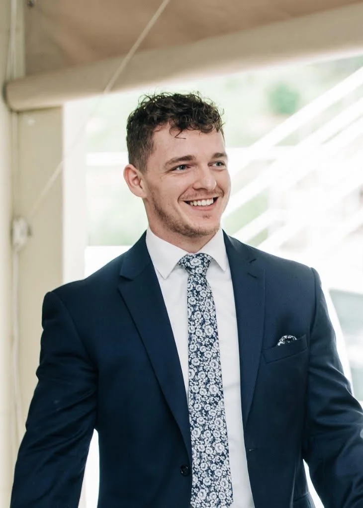A smiling man in a navy suit, white shirt, and patterned tie inside a bright space with large windows.