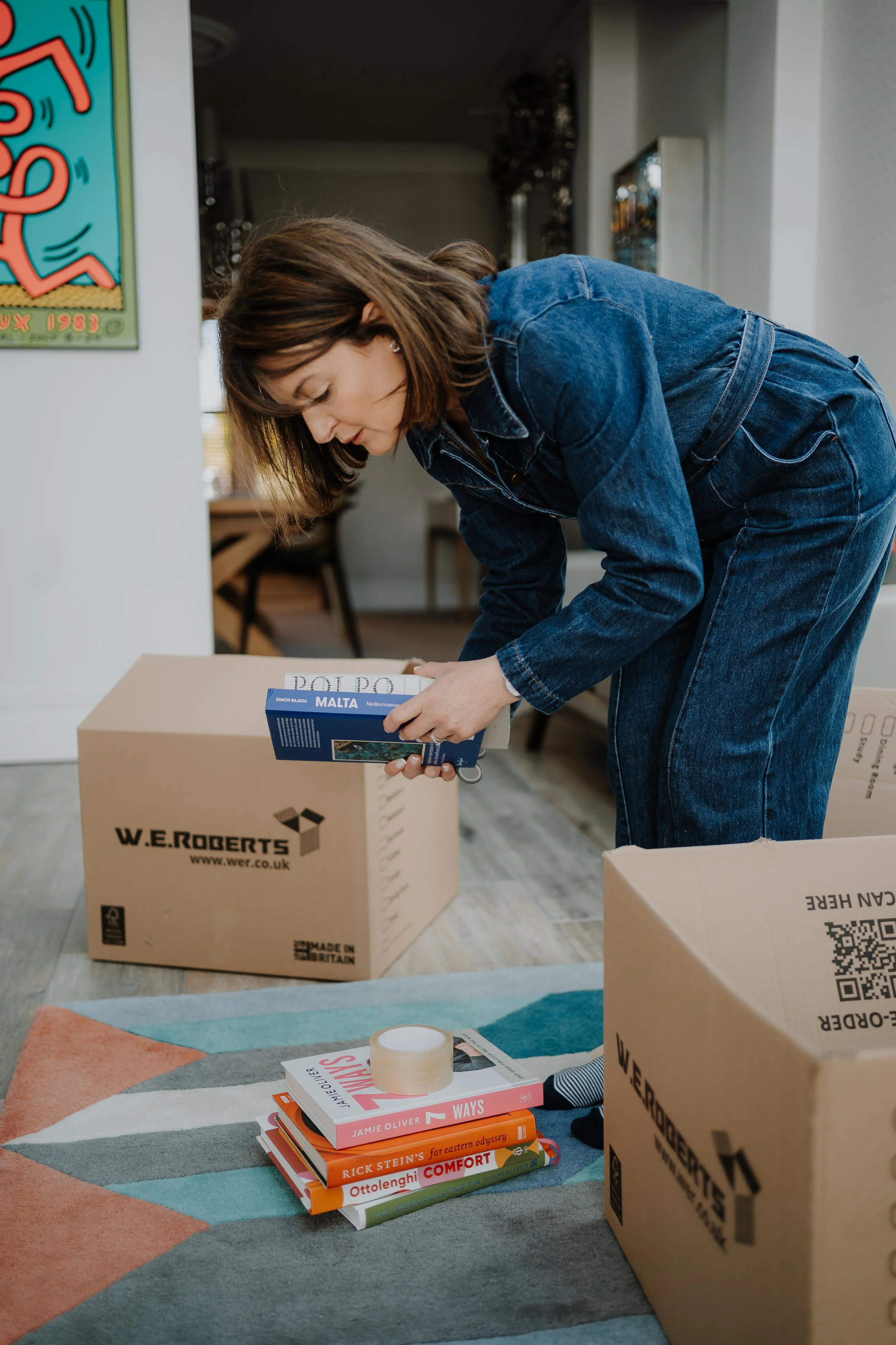 A woman in a denim outfit is bending over a cardboard box, with a stack of colorful books on a multicolored rug nearby. The books include titles by Jamie Oliver, Rick Stein, and Ottolenghi. There are additional cardboard boxes in the background, and the setting appears to be a home or an office space with bookshelves, furniture, and a window.