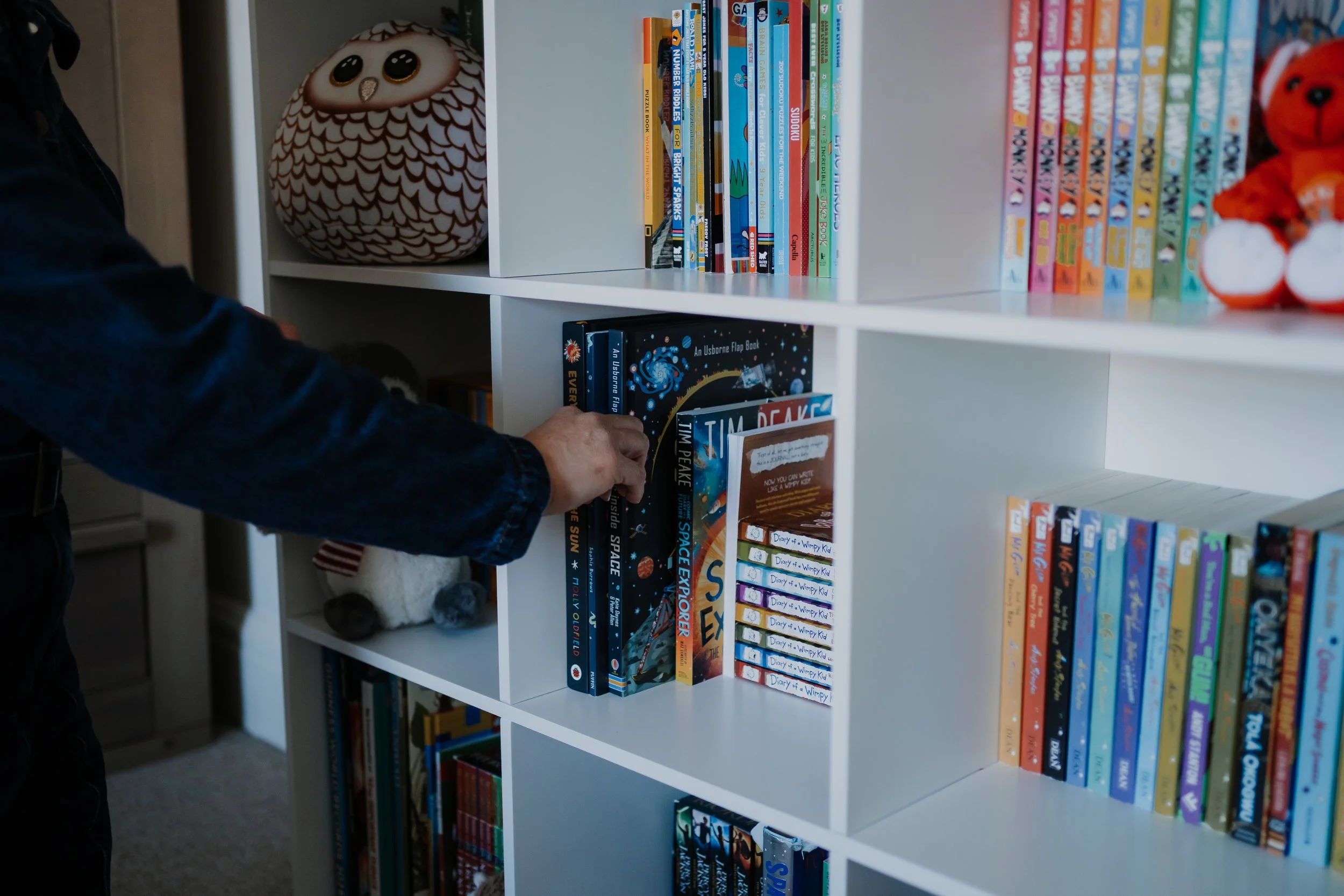 Part of a white bookshelf with colorful children's books and plush toys, including a brown and white owl and an orange bear, with a person's hand reaching for a book.