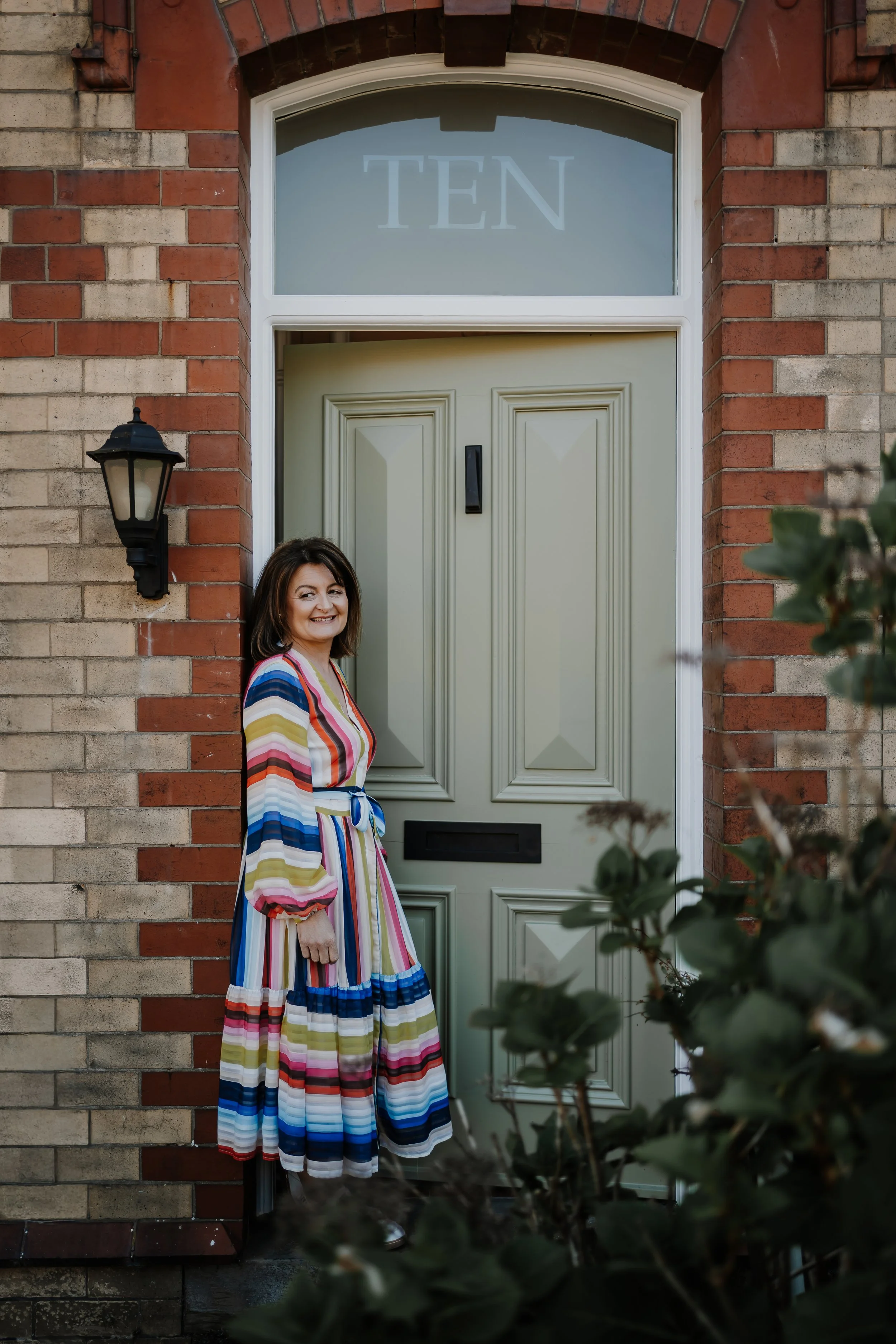 A smiling woman stands in front of a green front door with the number ten. She is wearing a colorful, striped dress and there is a black wall-mounted lantern on the brick wall beside her.