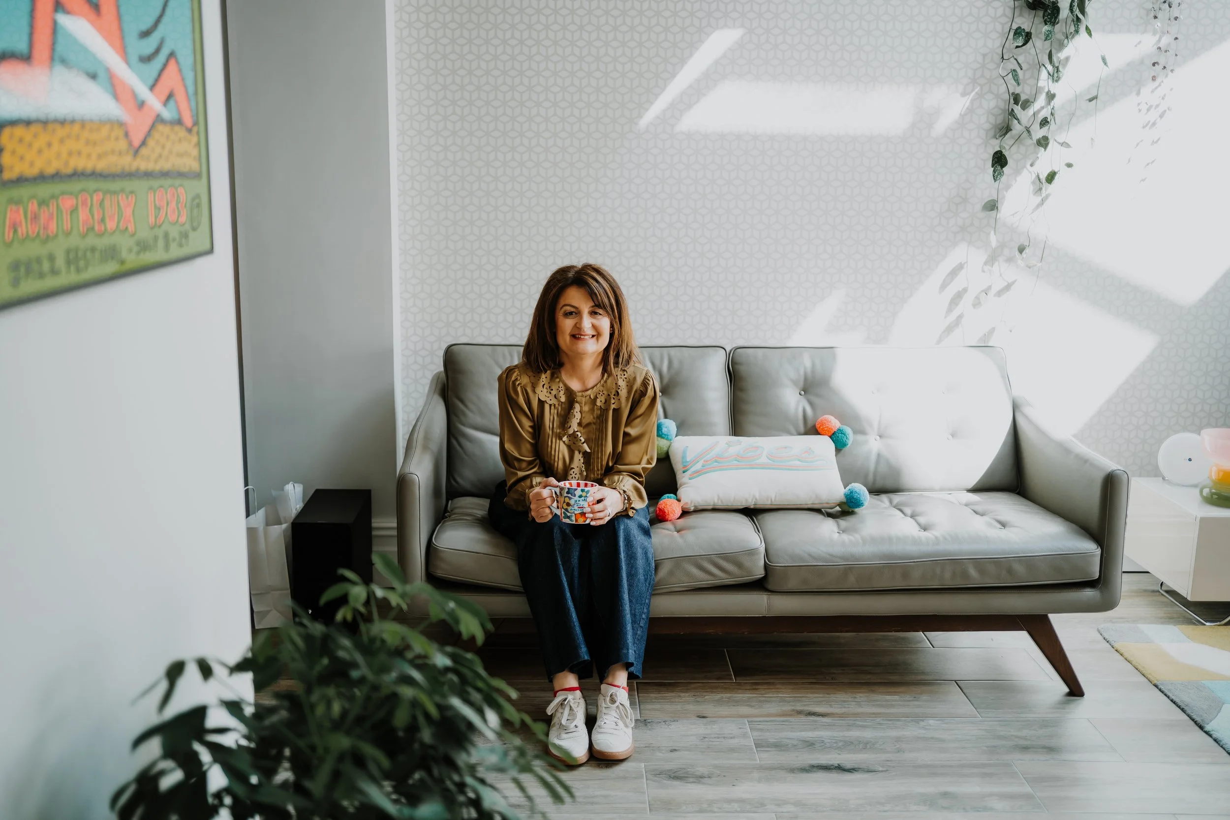 A woman with brown hair and a gold blouse sitting on a gray sofa, holding a colorful mug, smiling in a bright room with patterned wall, hanging plants, and decorated pillows.