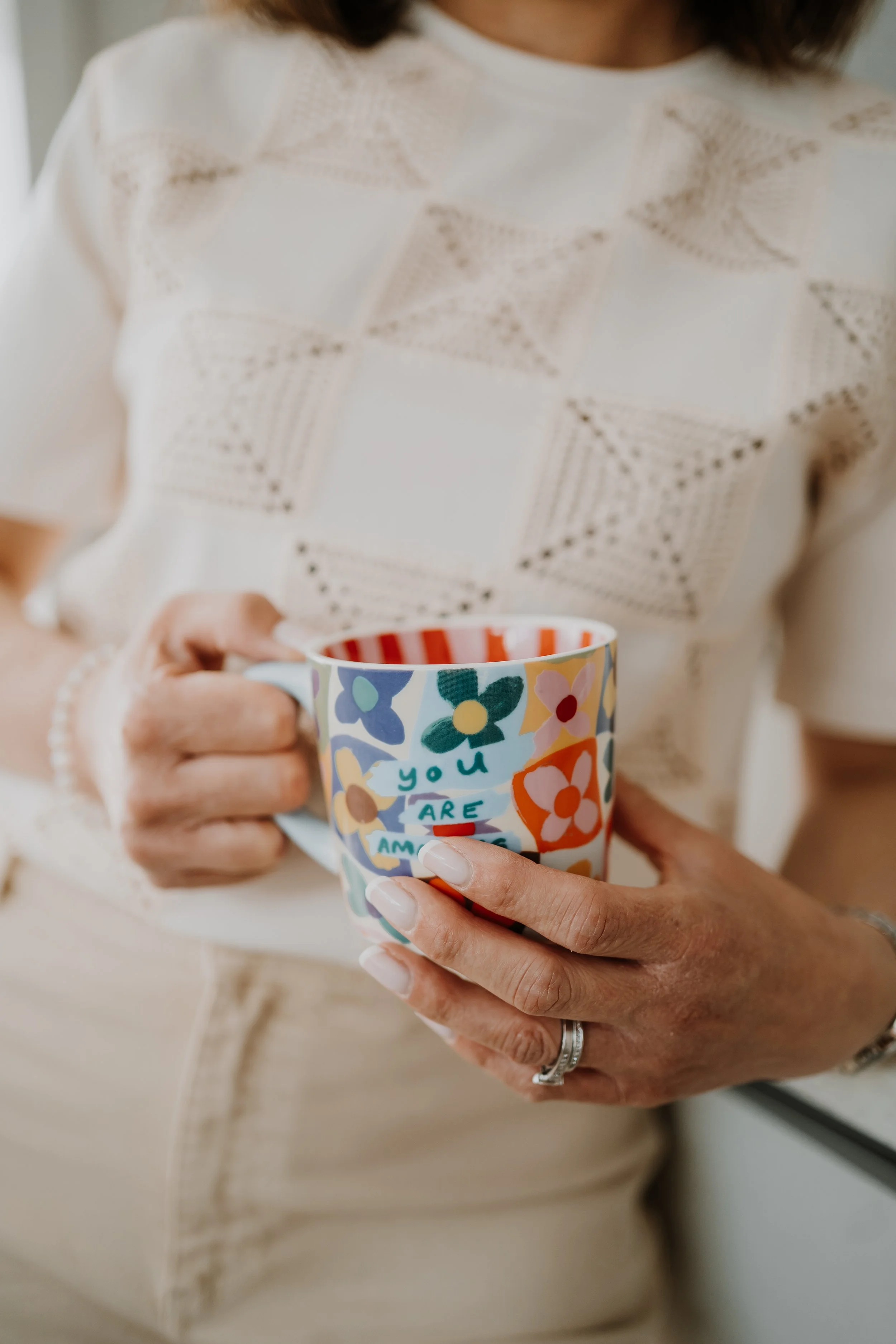 A woman holding a colorful coffee mug with a floral pattern and the words 'you are amazing' written on it.