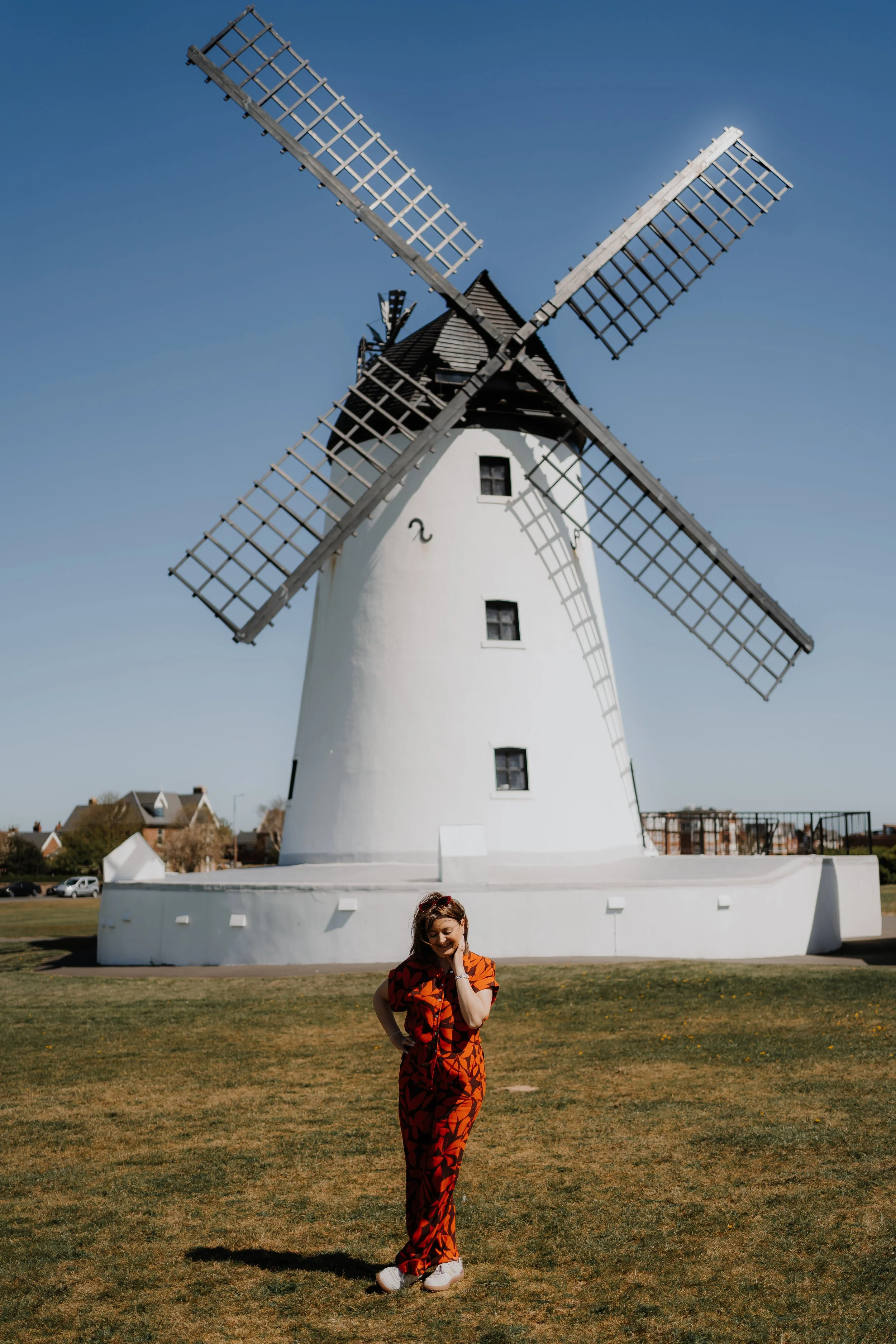 A woman in a red and black patterned jumpsuit standing on a grassy area in front of a white windmill with four large blades under a clear blue sky.