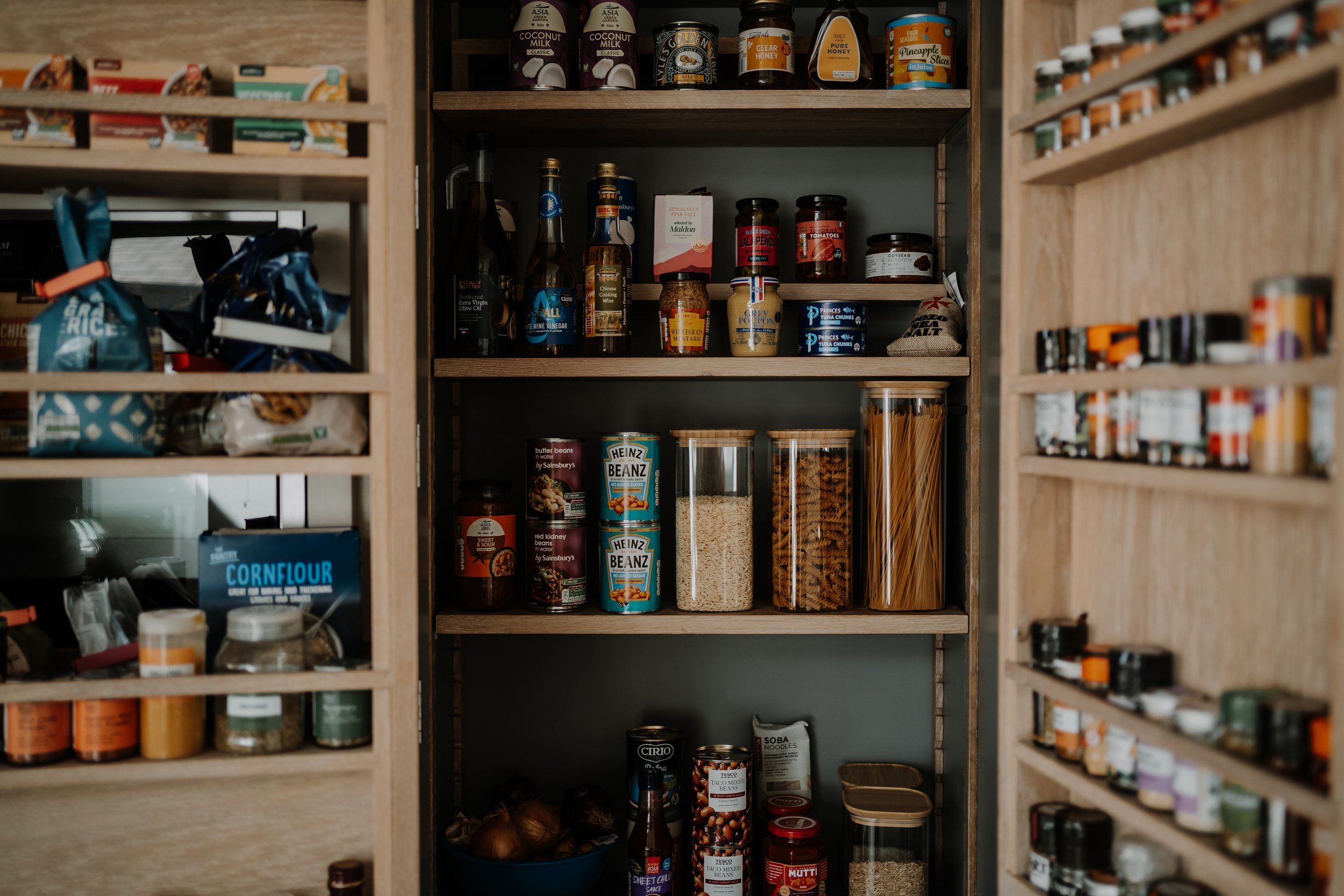 Pantry shelf with jars of spices, canned goods, pasta, sauces, and condiments, organized on wooden shelves.