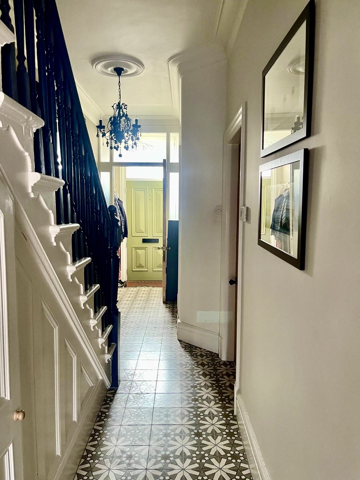 Hallway with patterned floor tiles, framed pictures on the wall, a staircase with white risers and dark wooden rails, a ceiling chandelier, and a green front door with glass panels.