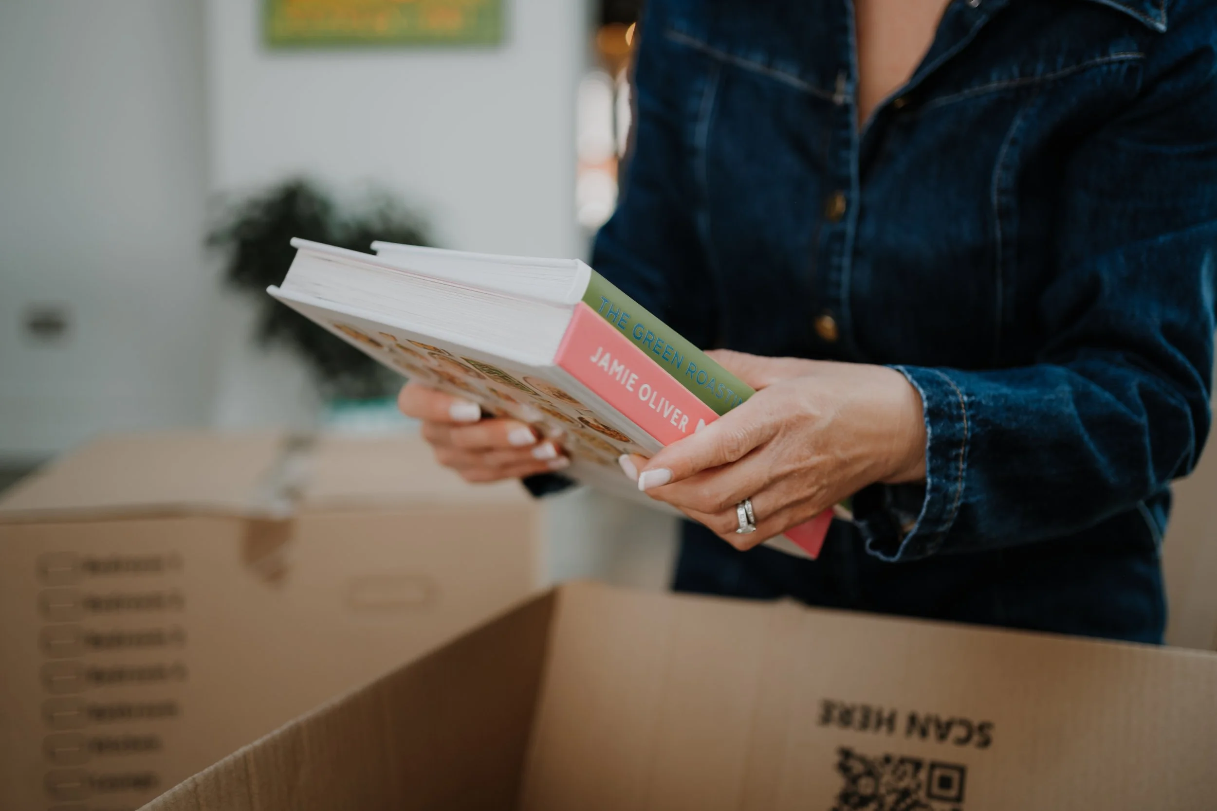 Person in denim shirt holding a stack of books in a room with cardboard boxes and green plant in the background.