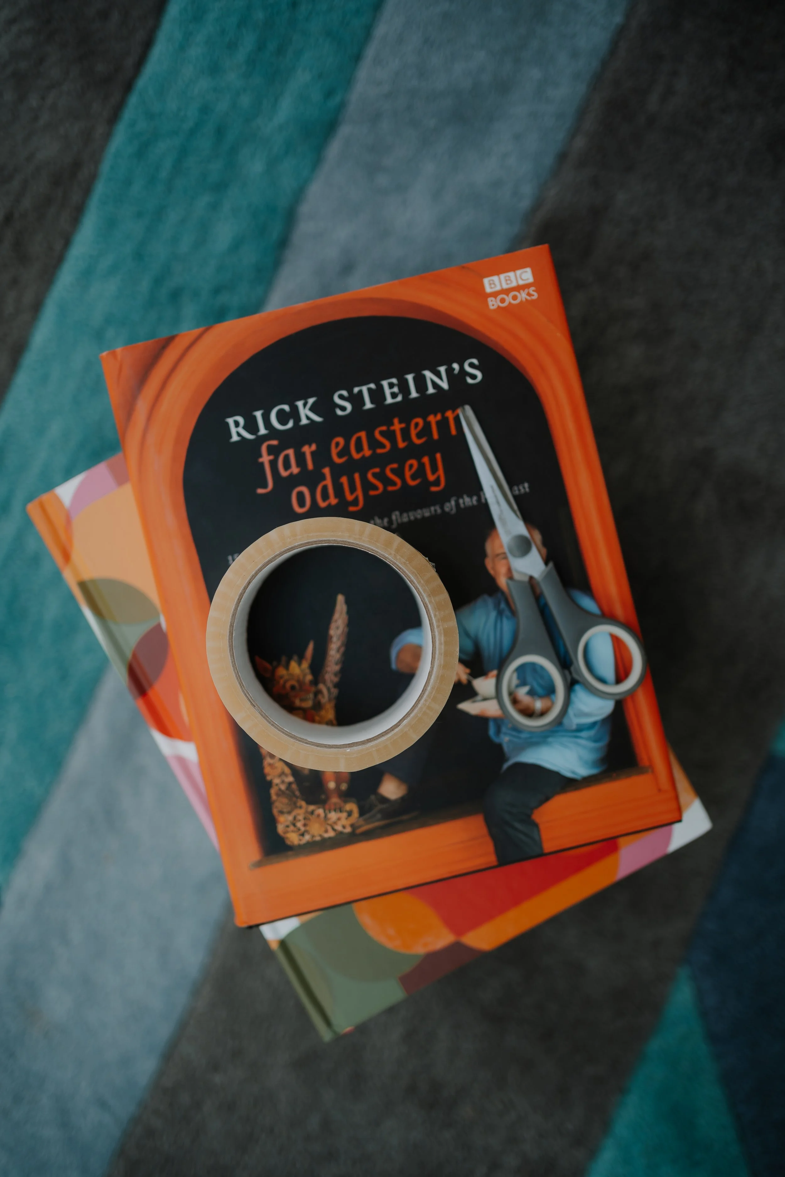 Close-up of a book titled "Rick Stein's Far Eastern Odyssey" resting on colorful magazines, with a roll of clear adhesive tape in the foreground.