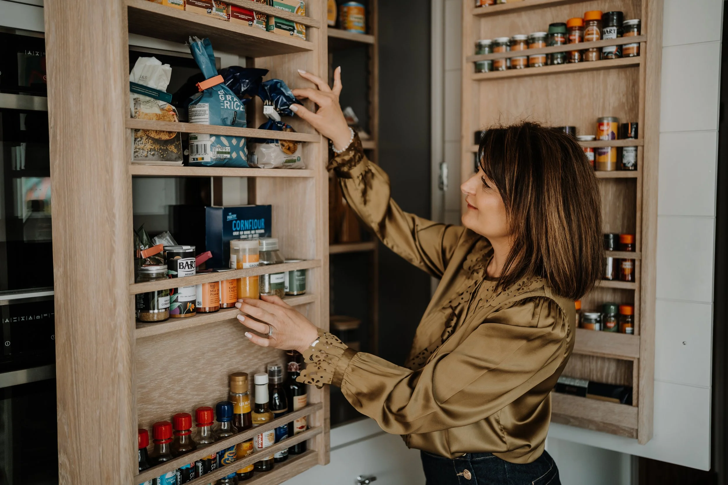 A woman with brown hair wearing a gold satin blouse stands in front of a wooden spice rack, reaching for a container of herbs or spices. She is smiling and appears to be selecting an ingredient from the shelf in her kitchen.