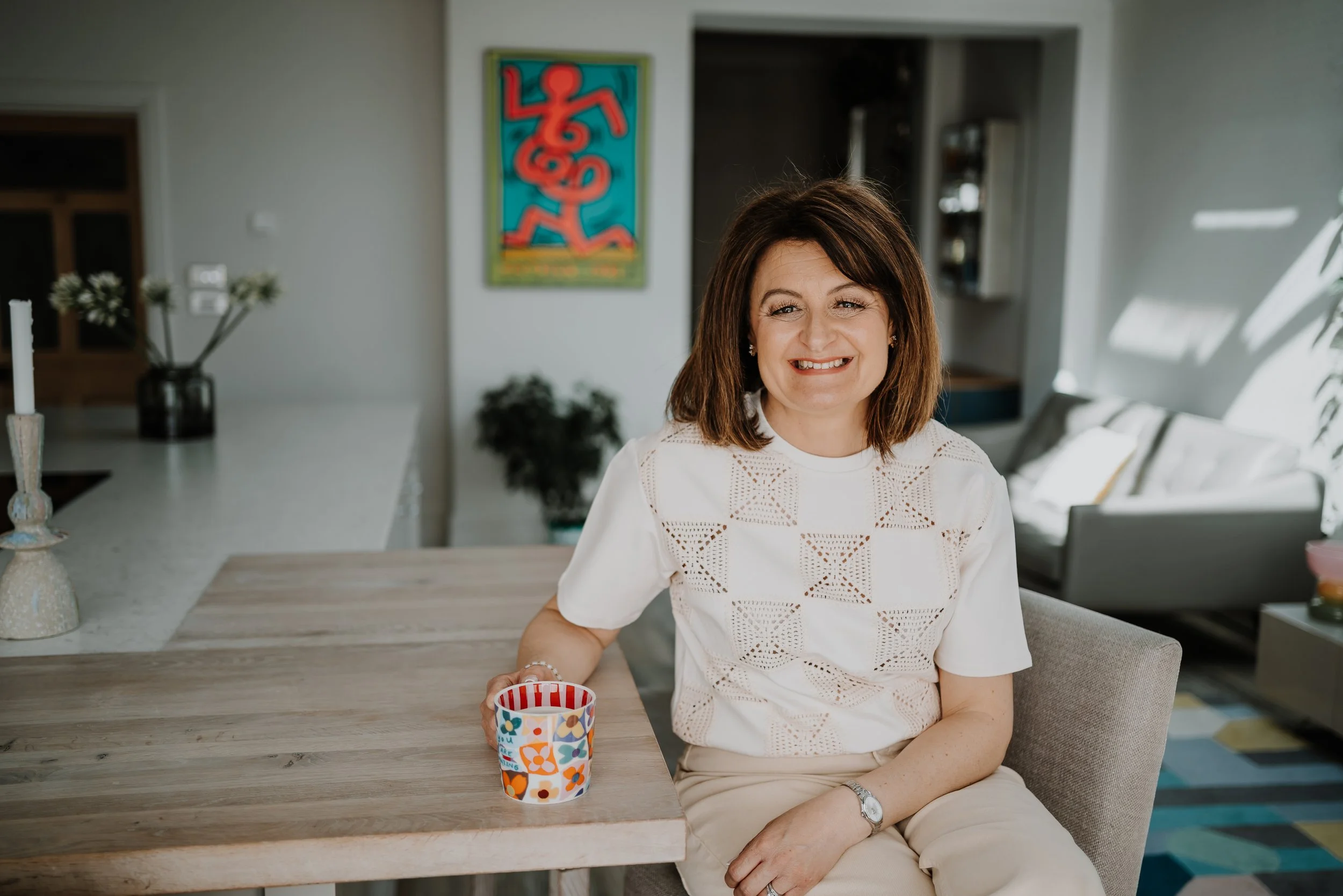 A smiling woman with shoulder-length brown hair, wearing a white lace blouse, sitting at a dining table with a colorful cup in front of her in a bright, modern home.