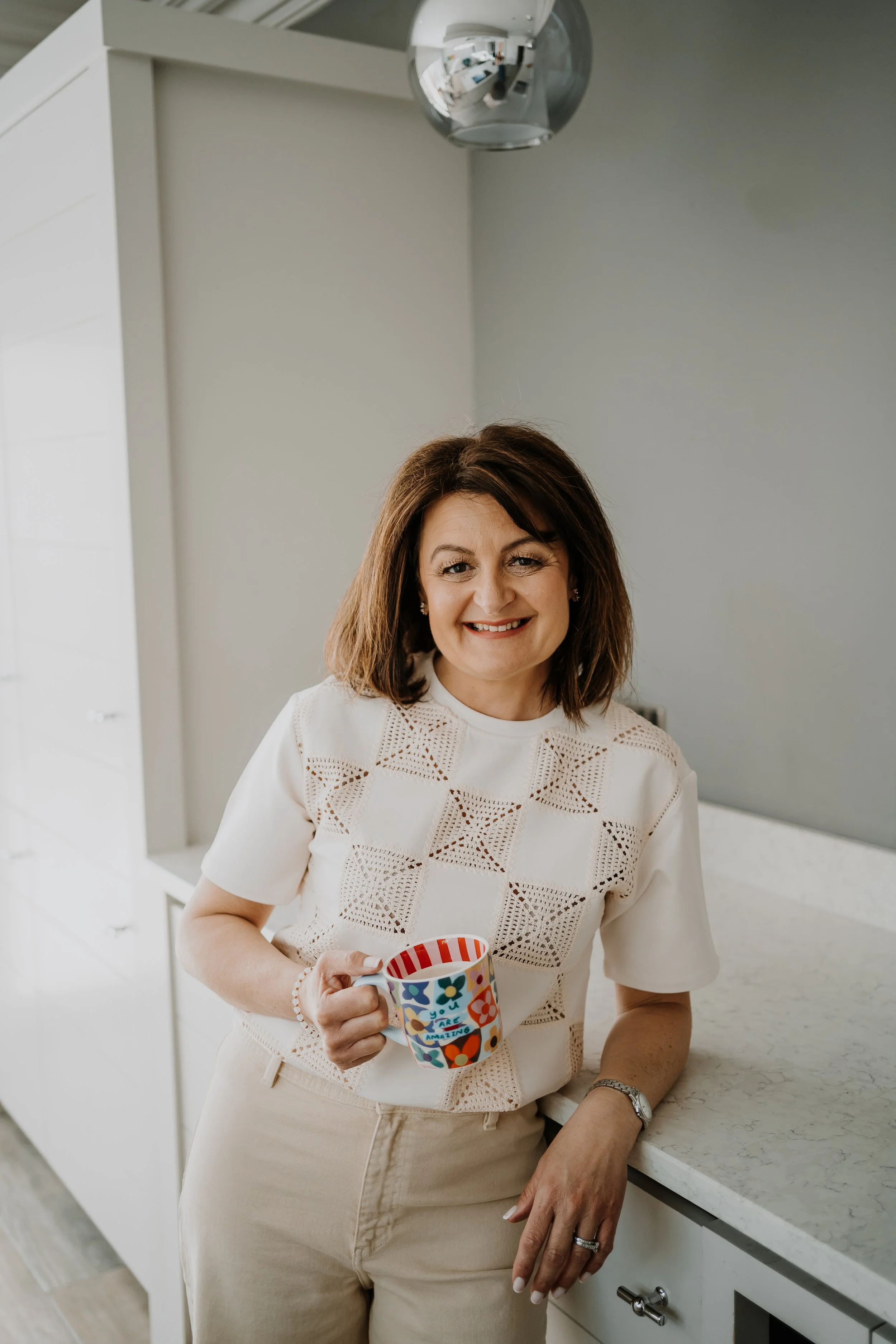 A woman with shoulder-length brown hair, smiling and holding a colorful coffee mug in a kitchen with white cabinets and a light-colored marble countertop.