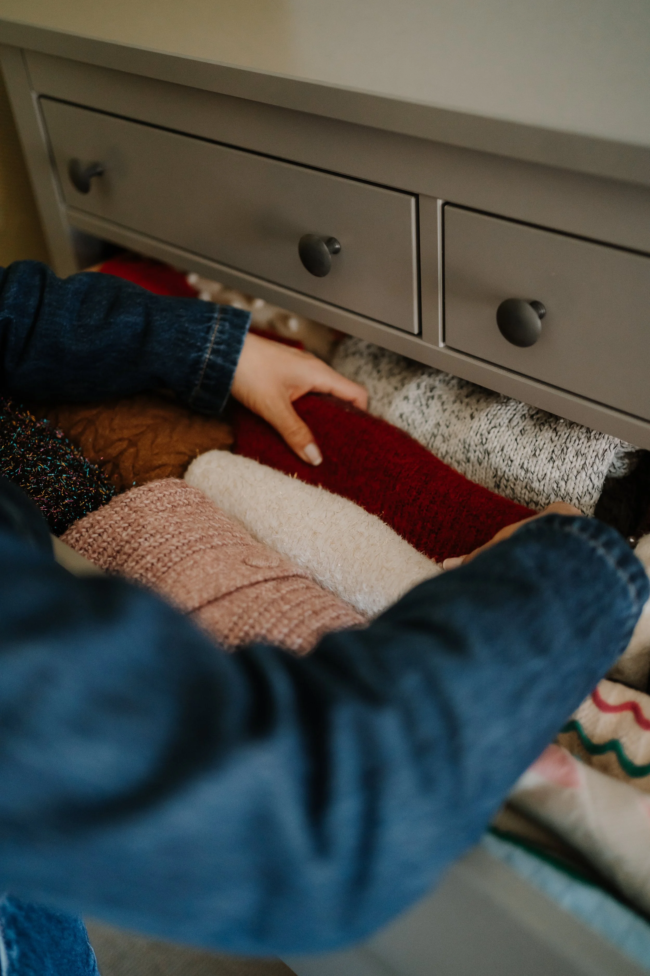 A person's hand reaching under a gray drawer chest, with various colorful blankets beneath. The drawer chest has round black knobs and appears to be in a cozy setting.