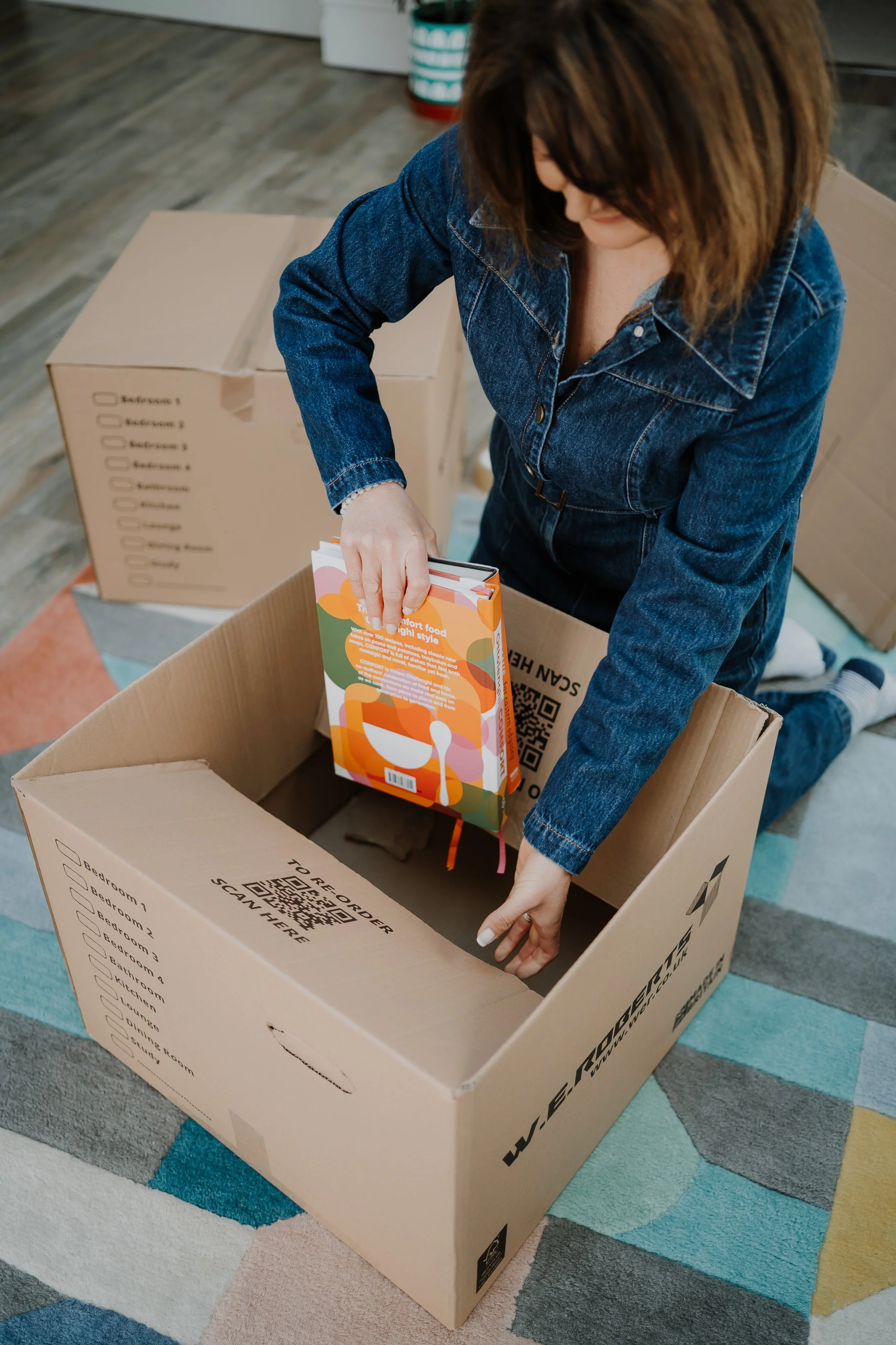 A woman in a denim jacket packing colorful food containers into a large moving box on a multicolored rug.
