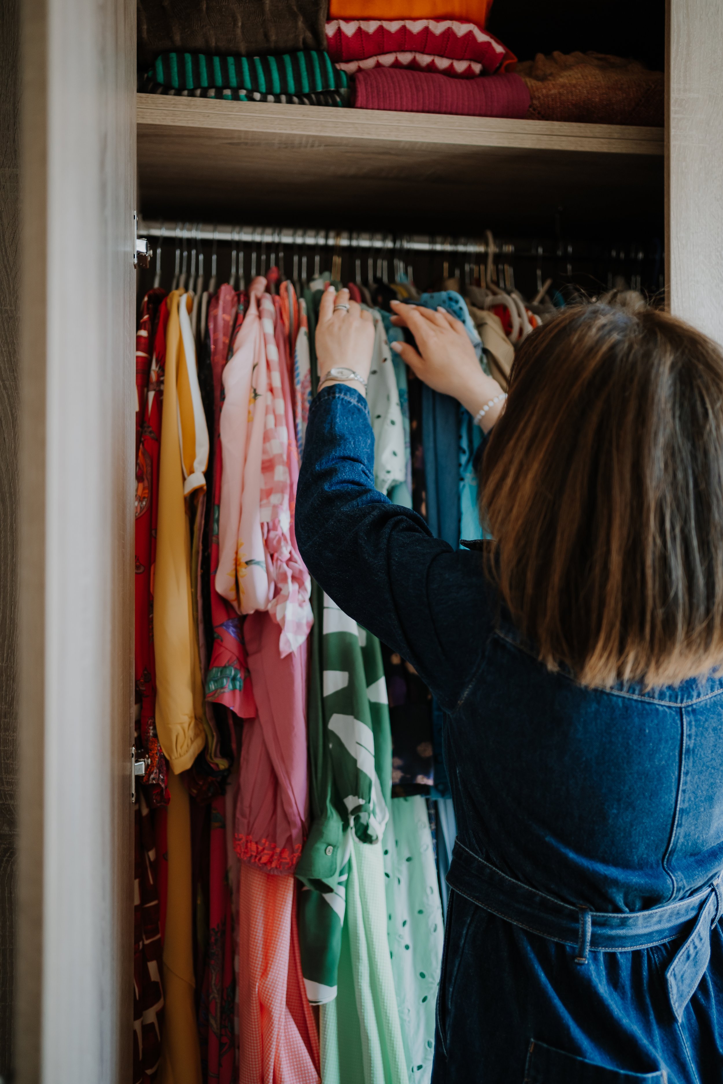 A woman is reaching for clothes in a closet, which contains colorful garments on hangers, including dresses and shirts, with folded clothing on a top shelf.
