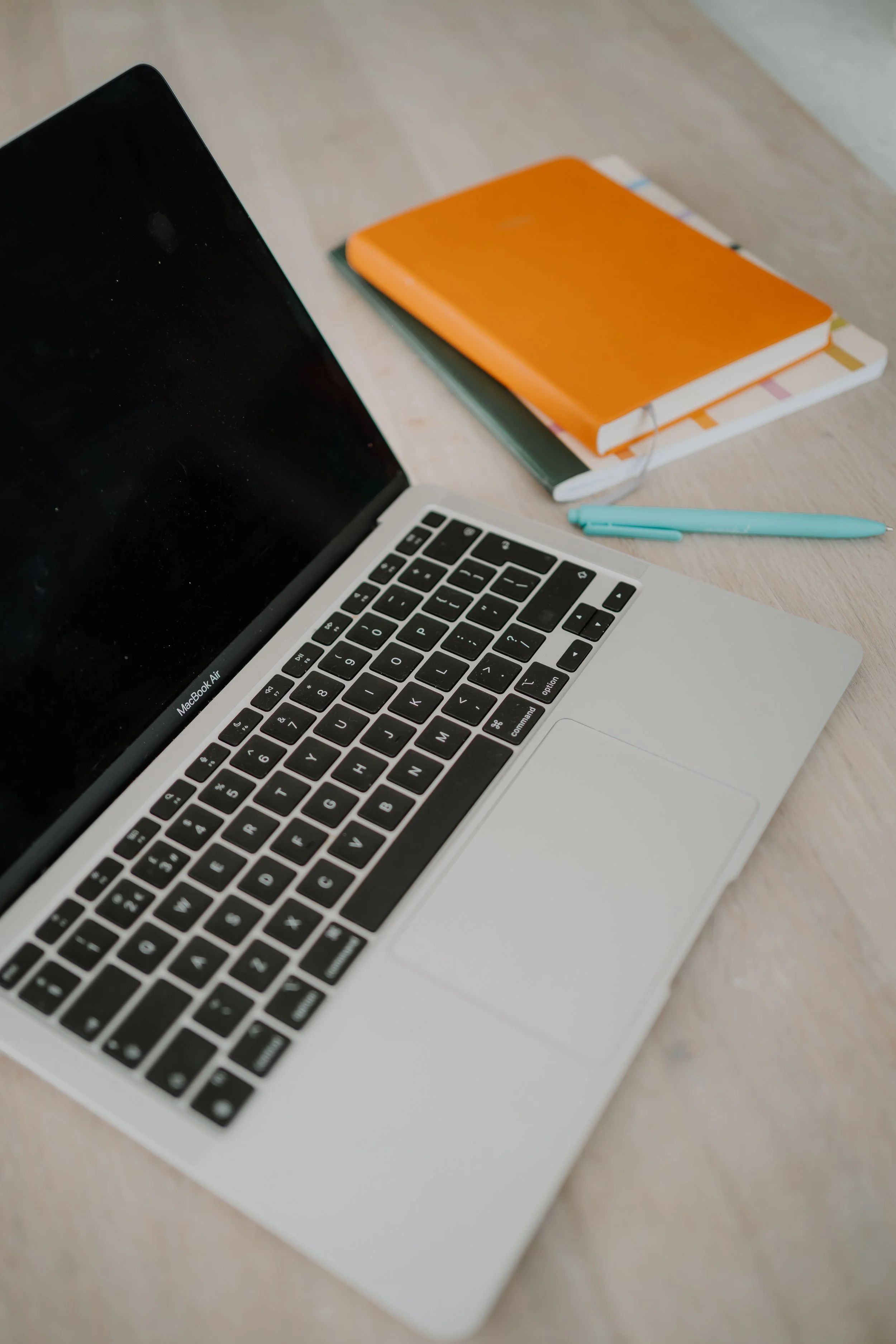 A laptop with a black screen, a blue pen, and two notebooks, one orange and one with colorful branding, on a light wooden surface.